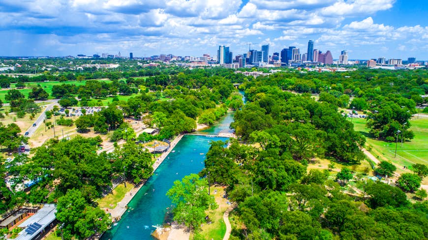 Aerial view of the Barton Springs pool, which is surrounded by tall green trees. In the distance, you can see the Austin skyline; Austin vs. Norman