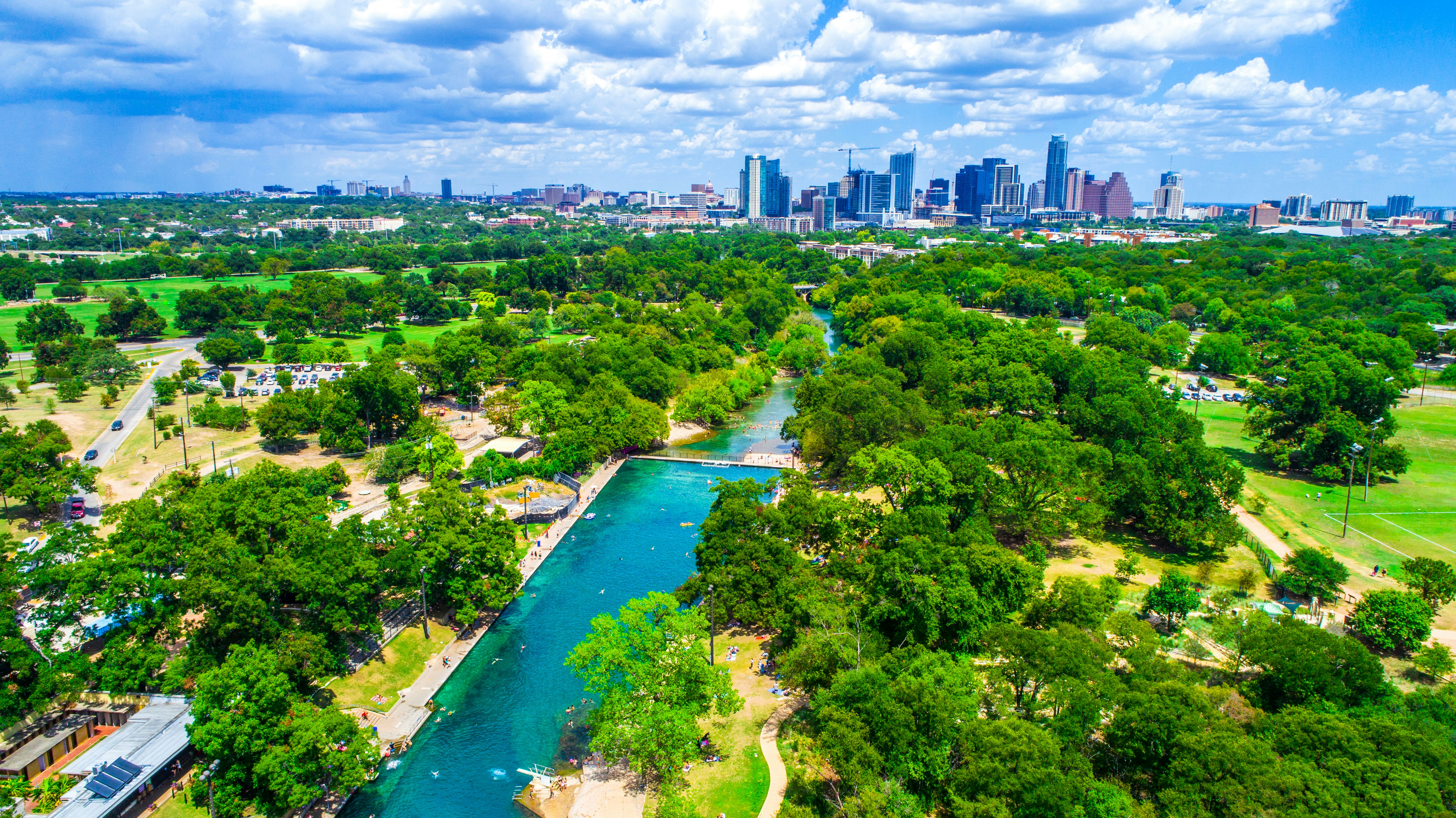 Aerial view of the Barton Springs pool, which is surrounded by tall green trees. In the distance, you can see the Austin skyline; Austin vs. Norman