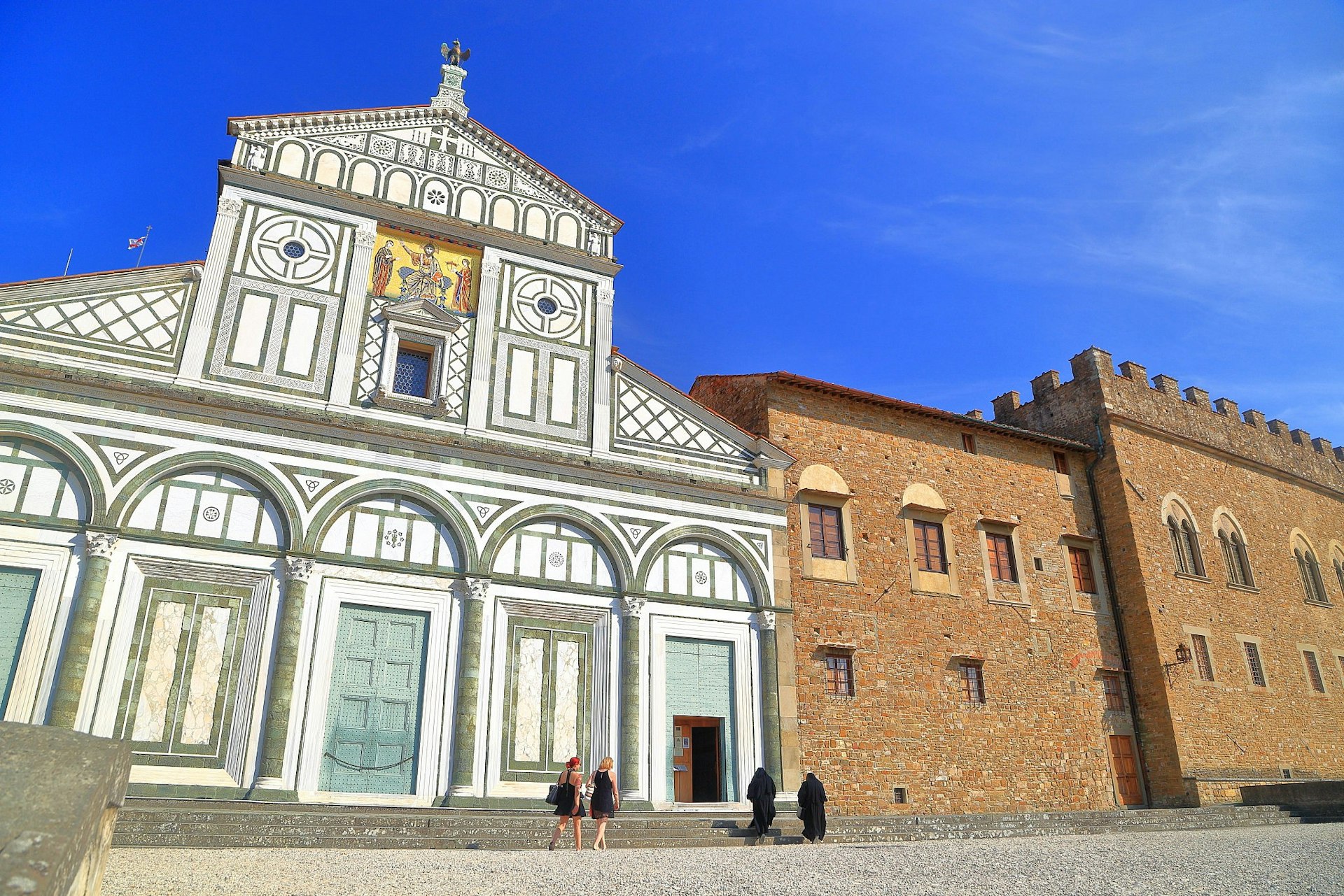 Two pairs of women - visitors in black dresses and nuns in habits - walking outside Florence's Basilica di San Miniato al Monte Florence, a church with an intricate multicoloured marble facade. 