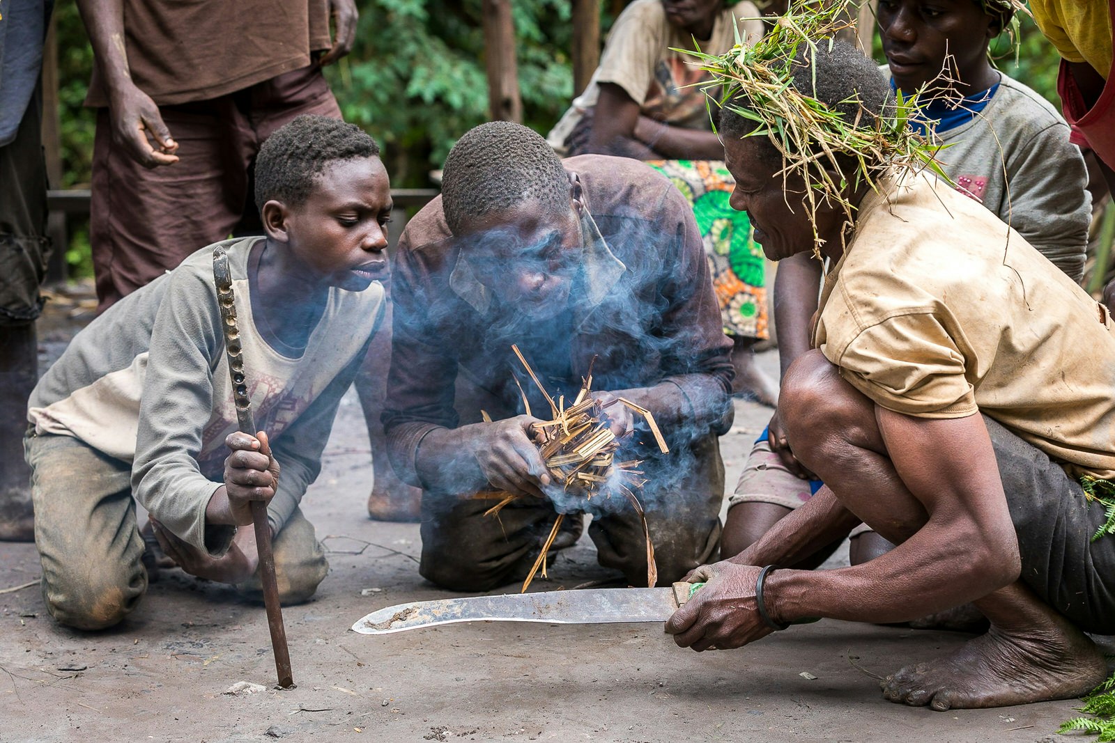 A kneeling Batwa man blows on a collection of dry grass in his hands that smokes; to his left is a boy holding the stick that was used to create the initial spark by spinning.