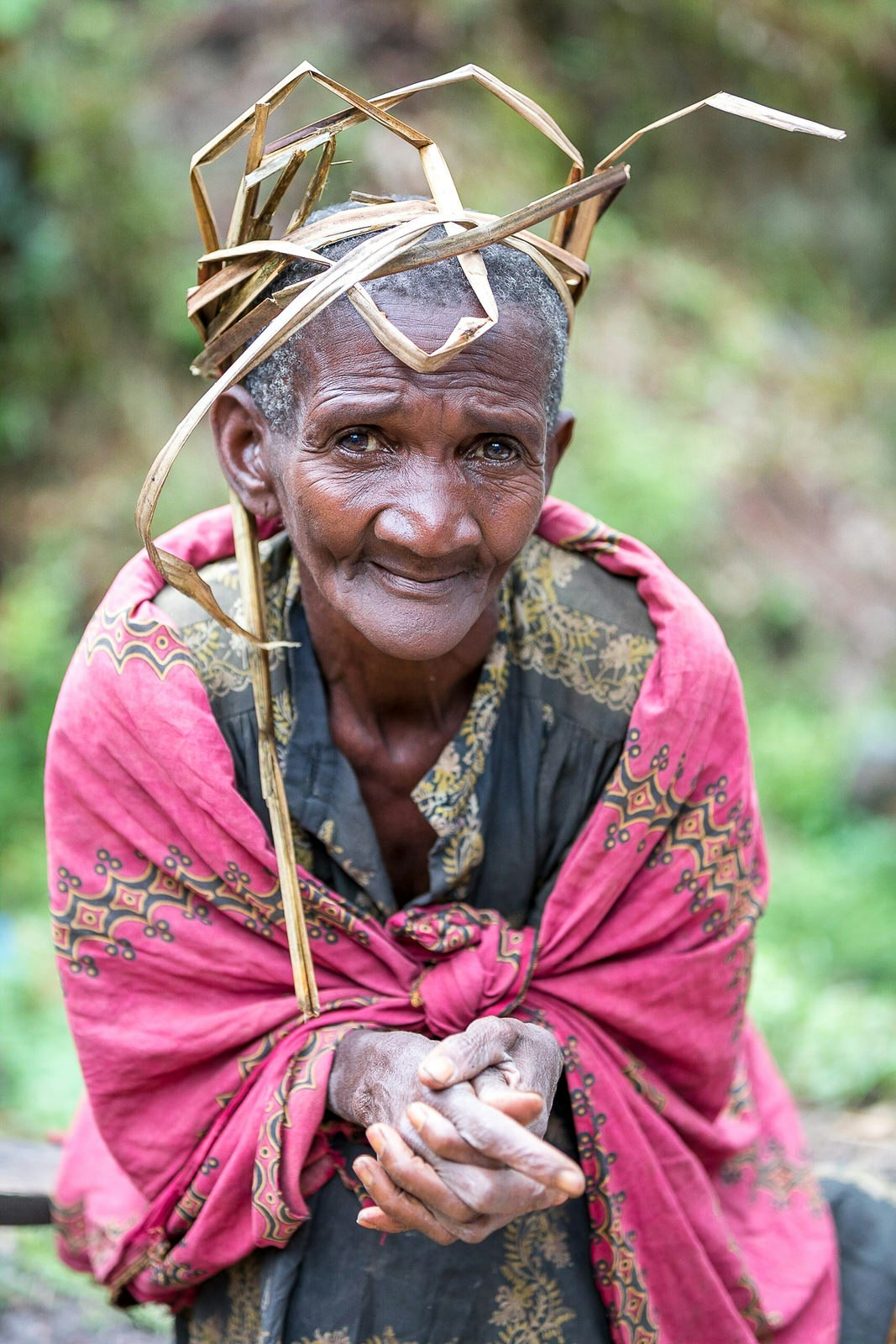 An elder Batwa wearing a pink blanket with yellow and black highlights leans over with a hunched back with her hands clasped in front of her; She has a head dress of long grass.