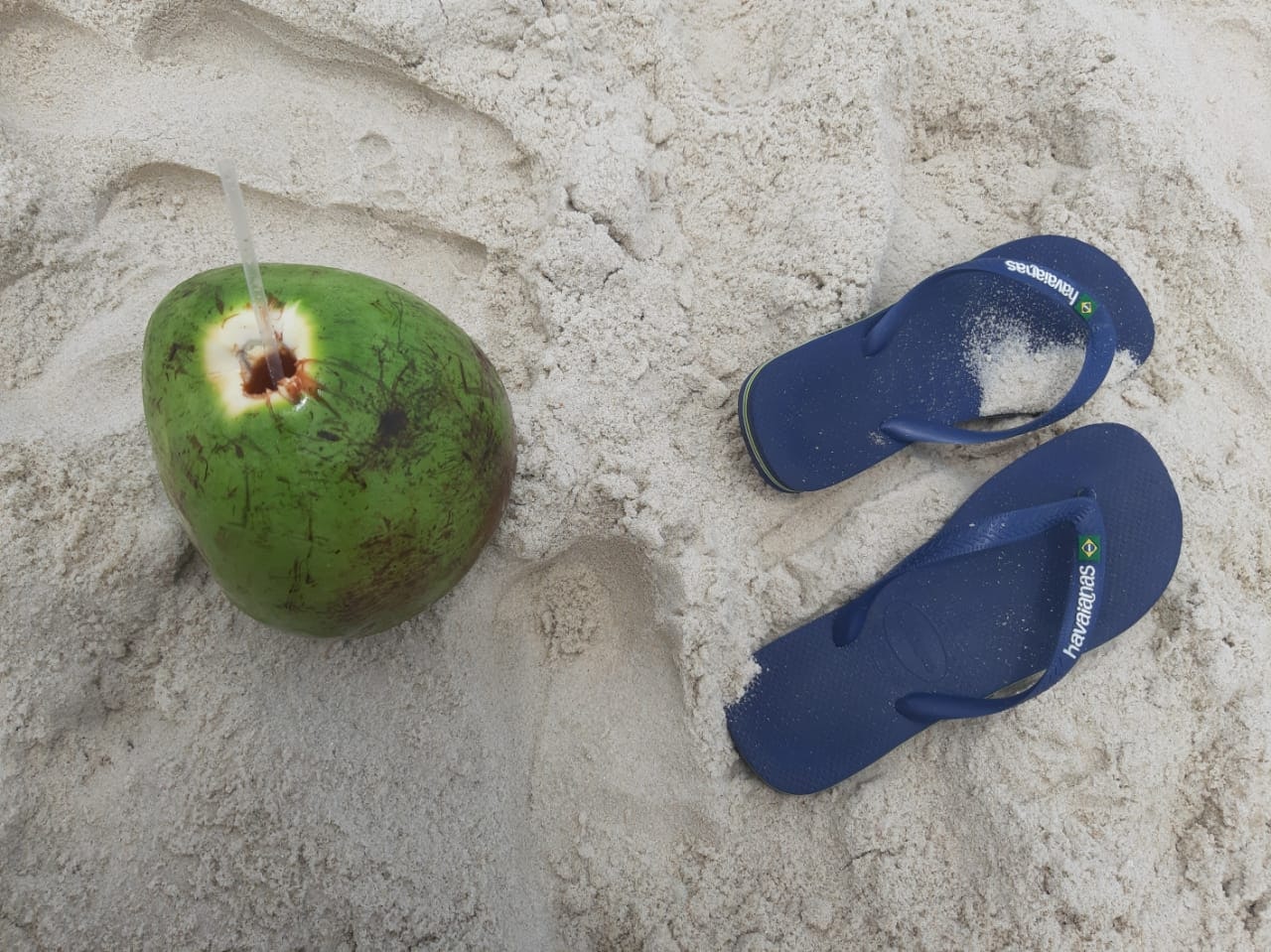 A coconut with a straw in it and a pair of flip flops on the sand