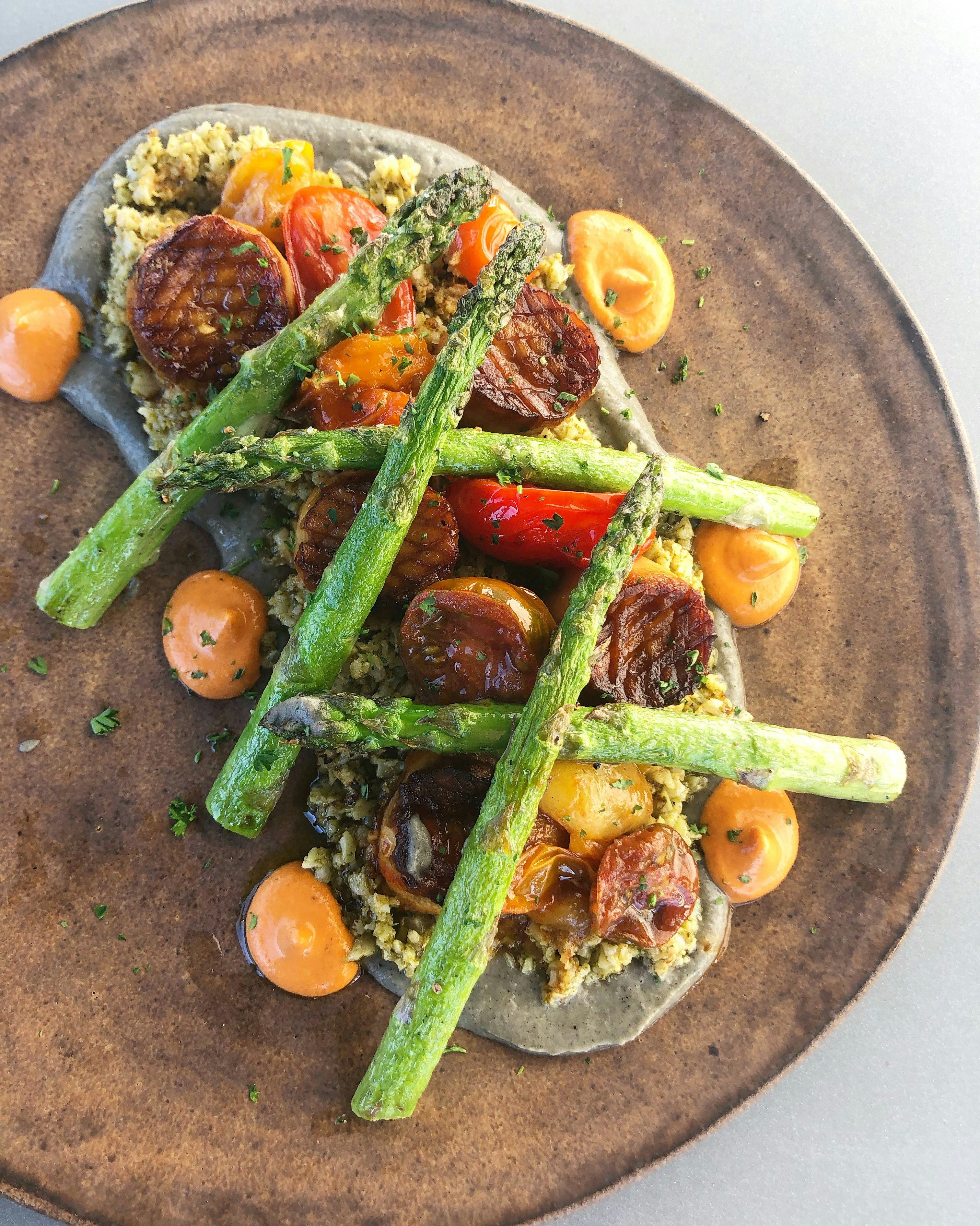 Medallion-sized trumpet mushrooms lay on a bed of hummus and cauliflower and topped with asparagus and tomatoes 