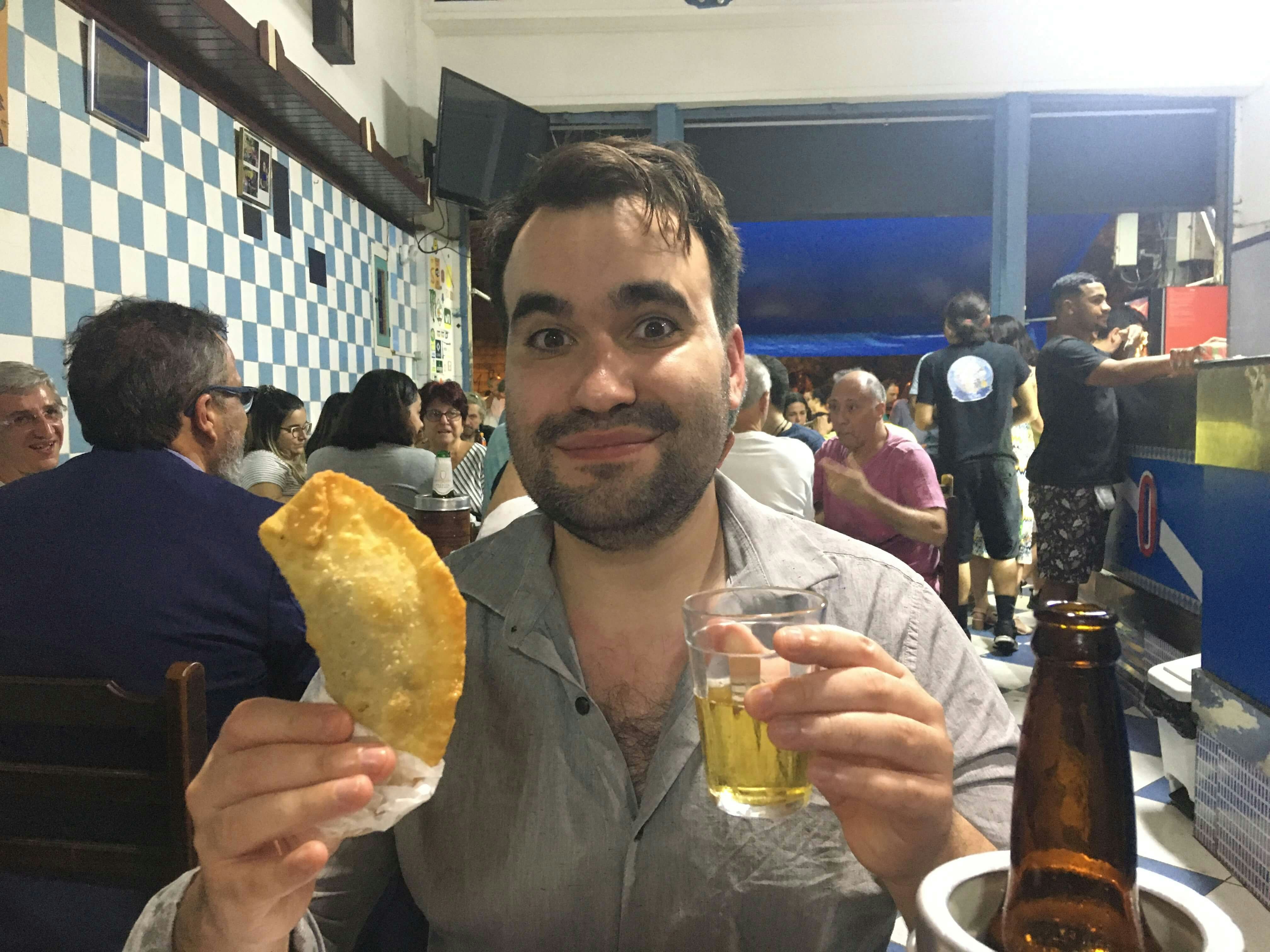 The author holds a glass of beer and a fried stuff pie. A busy restaurant is behind him
