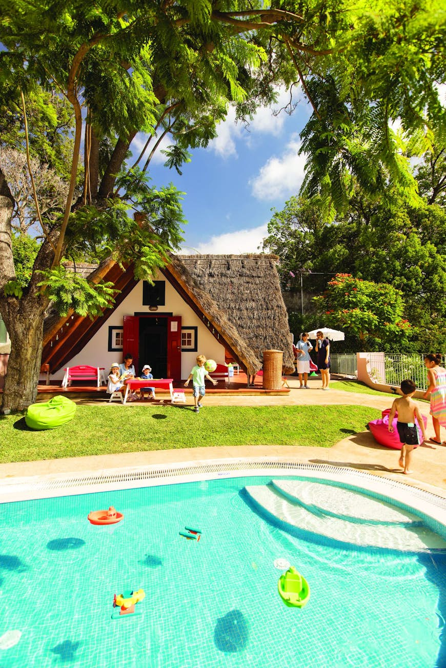 A small, thatched house by a pool at Belmond Reid's Palace, Portugal, pictured on a sunny day. A family can be seen sitting outside. 