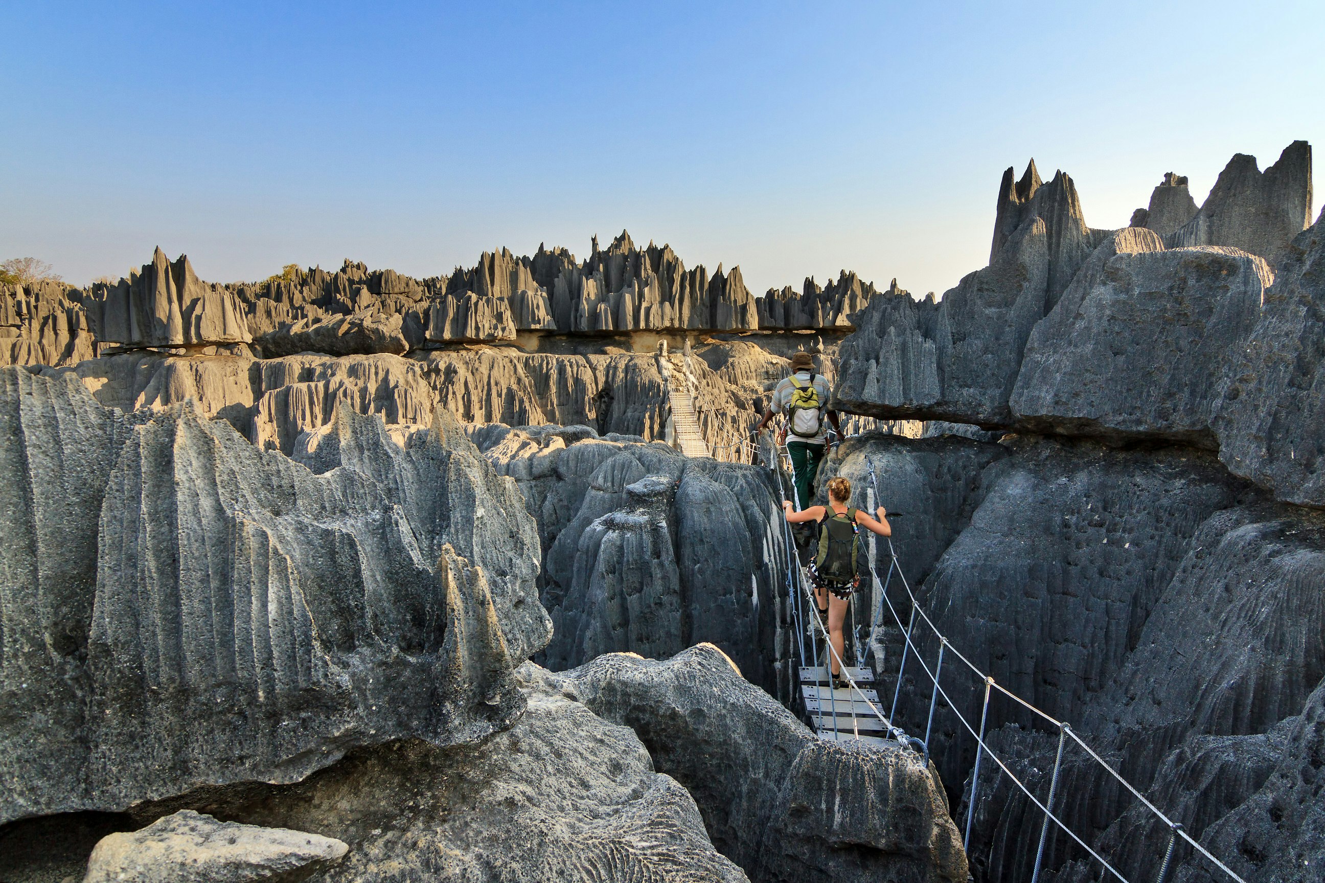 Two hikers make their way across a rope bridge that swings between the razor-like pinnacles atop the tsingy rock formations.