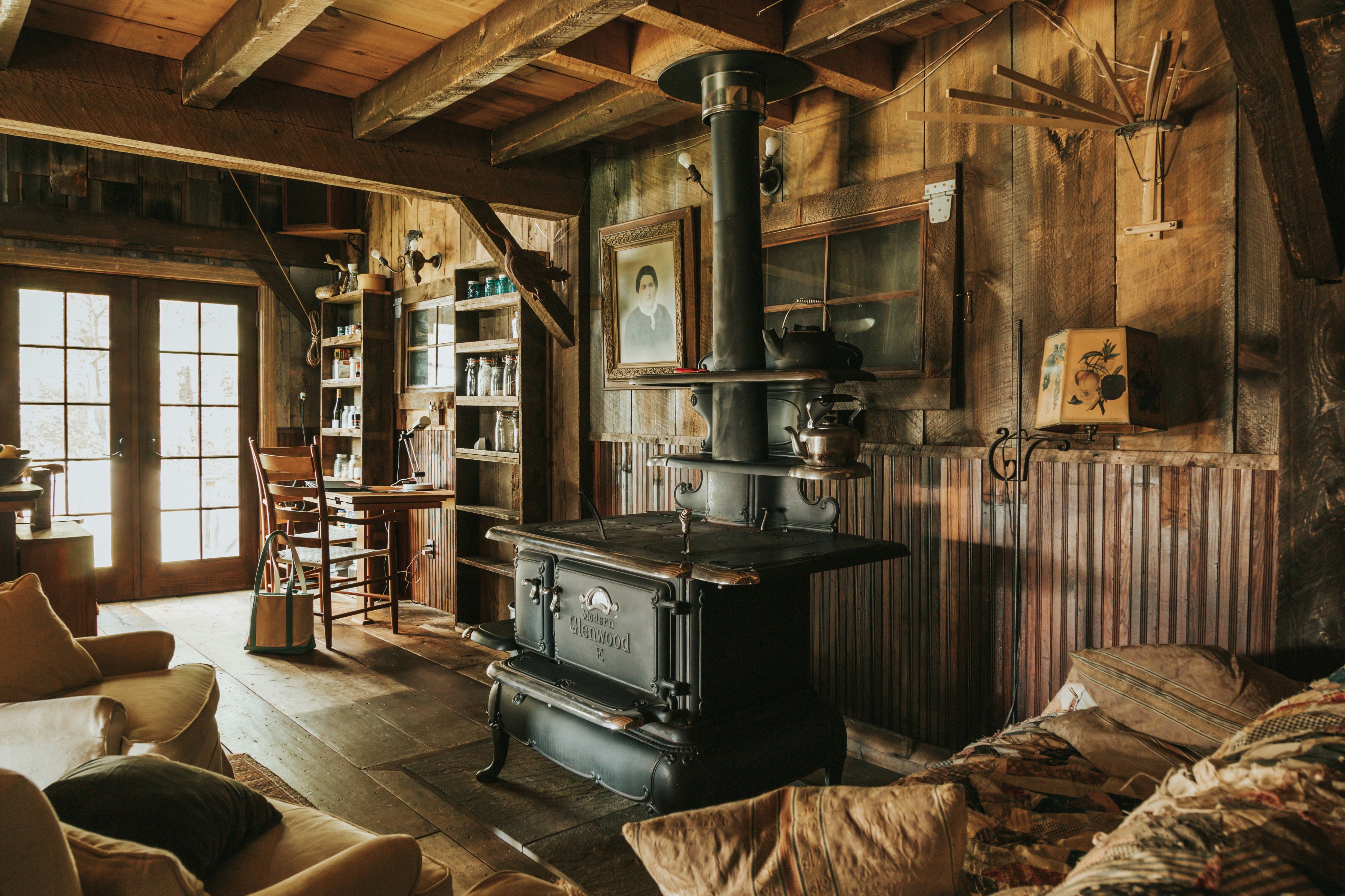 The inside of a cosy, wood-panelled cabin. The main focal point of the room is a large, black, cast iron range, and comfy chairs are arranged around it.