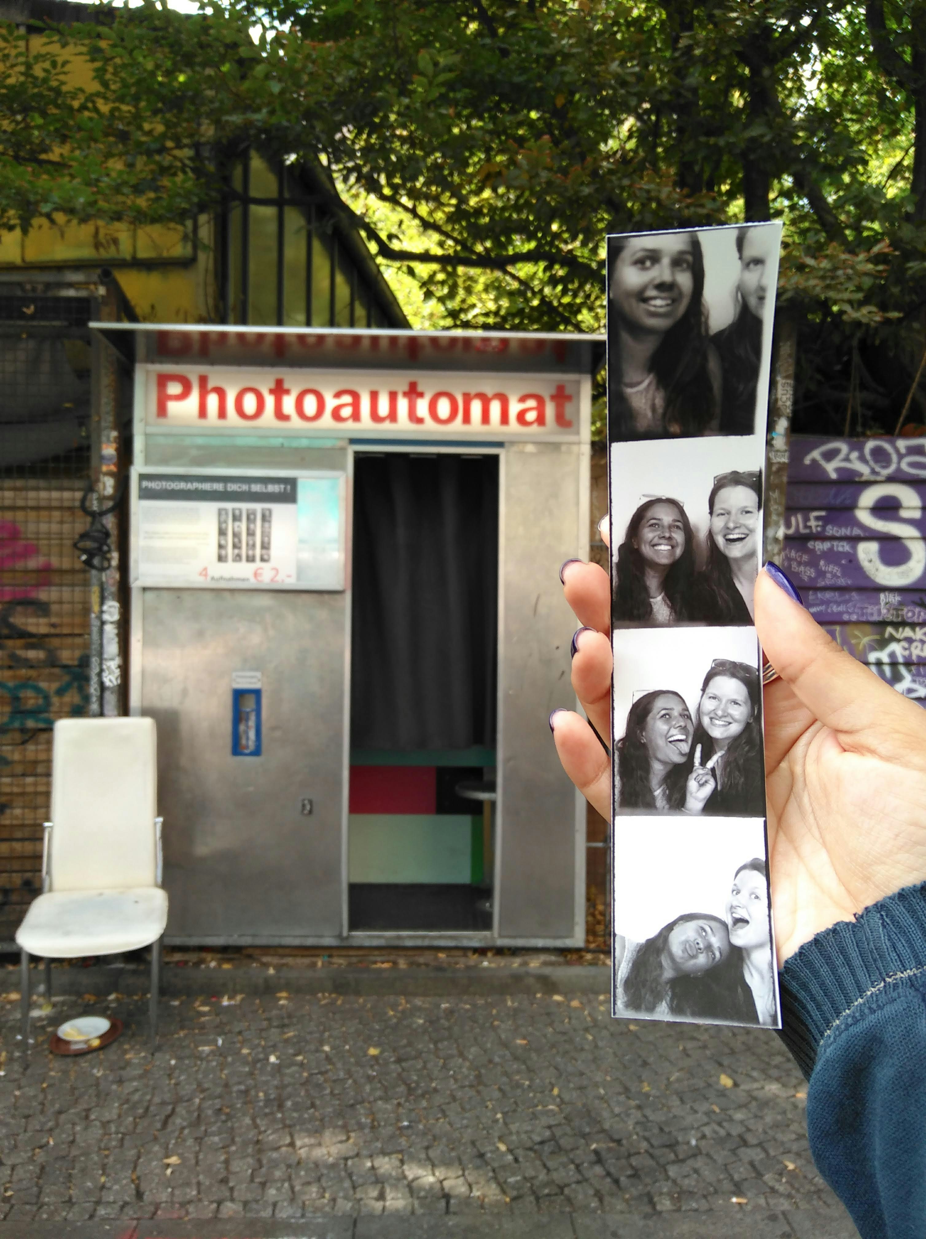 A Berlin 'Photoautomat' photo booth is in the background, while a hand reaches into the shot, holding a strip of four black-and-white photos of the writer and her friend.