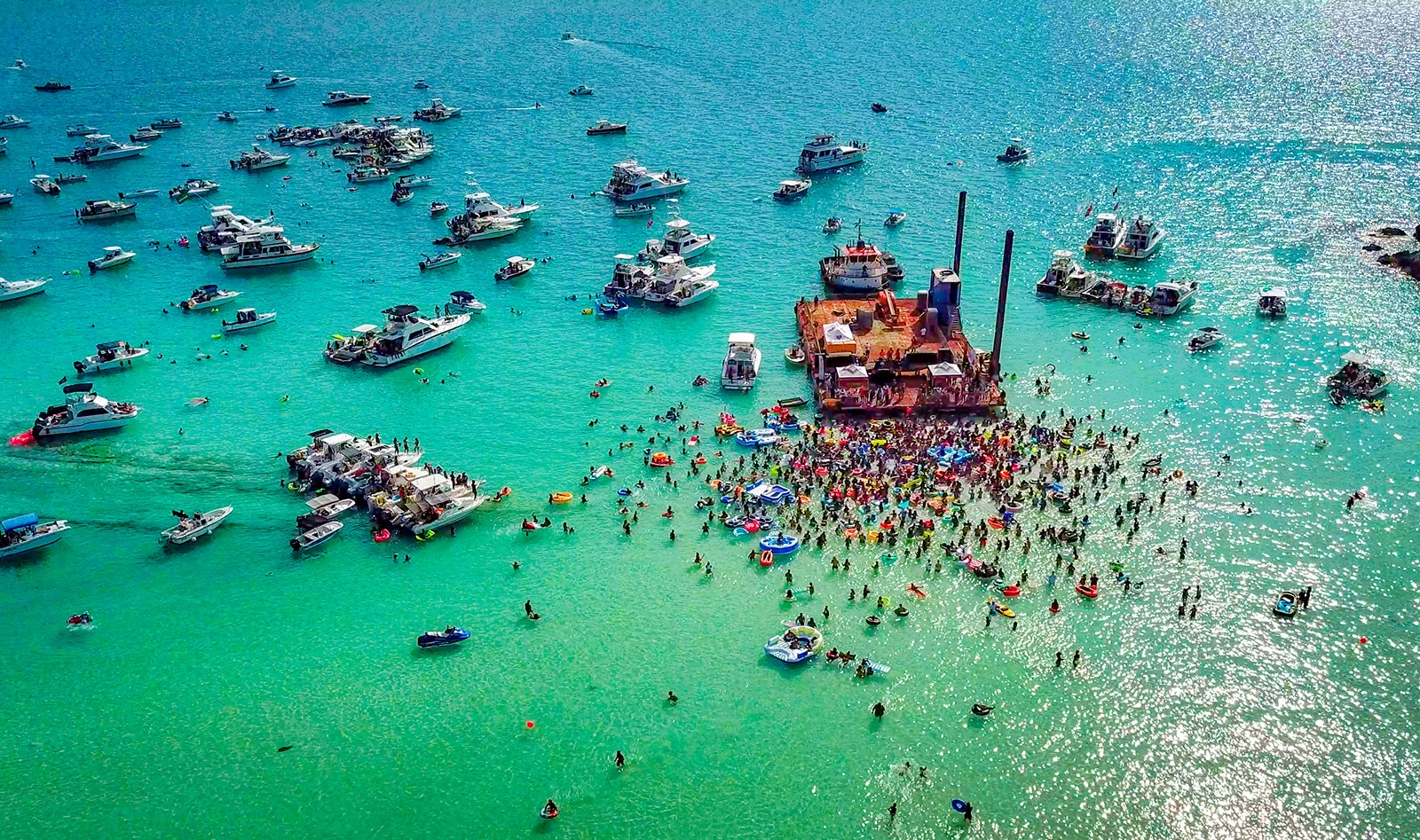 An aerial shot of boats and people in the water surrounding a large wooden raft