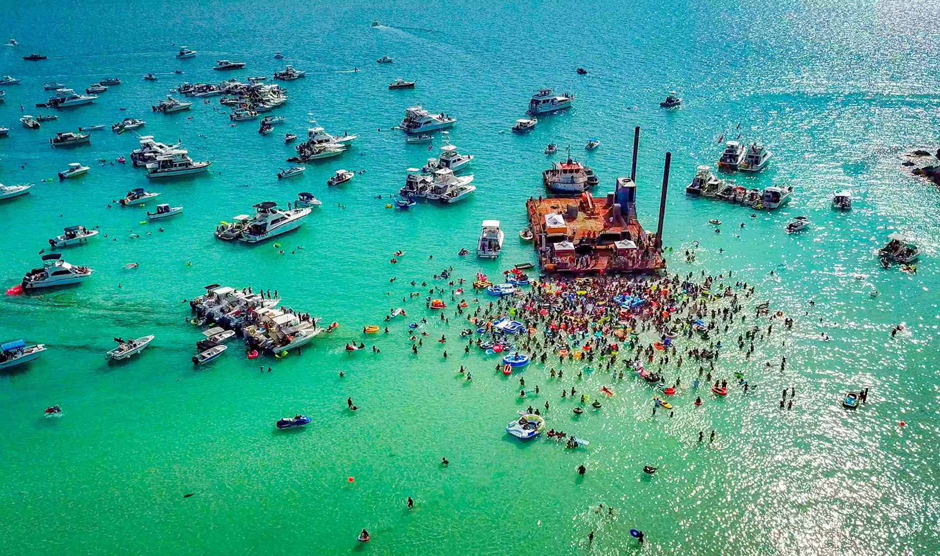 An aerial shot of boats and people in the water surrounding a large wooden raft