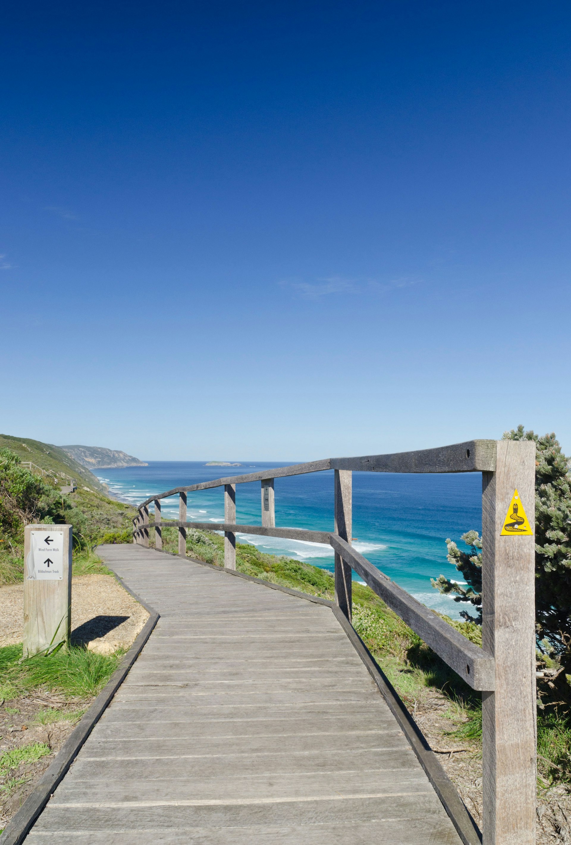 A boardwalk leads along a stunning section of coast in Western Australia; a yellow triangle badge on the railing has a black snake emblem, the sign for the Bibbulman Track.