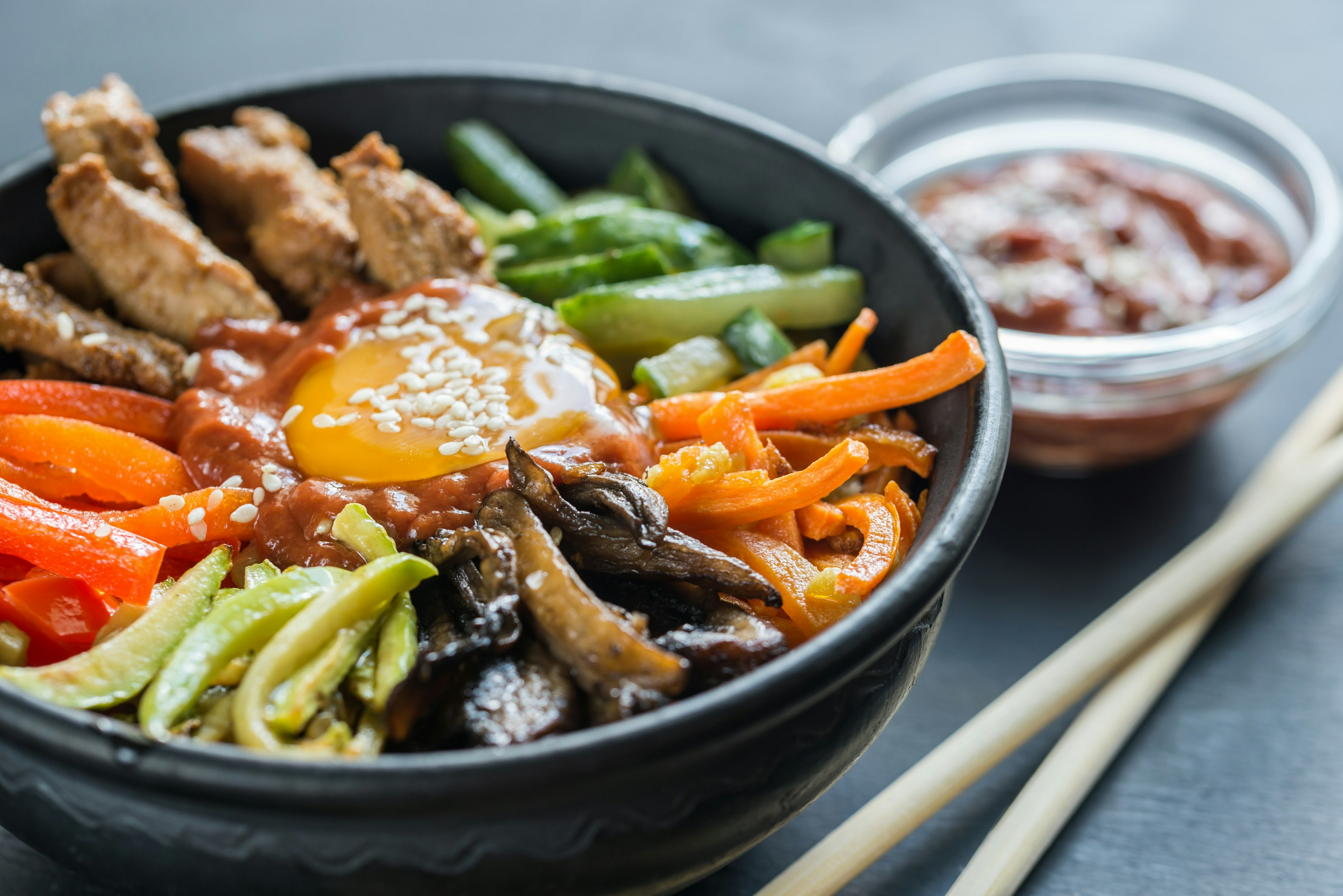 A close-up view of a bowl of bibimbap, a Korean rice dish which also contains vegetables and shredded meat. Alongside the black bowl containing the food is a small bowl of sauce and a pair of chopsticks.