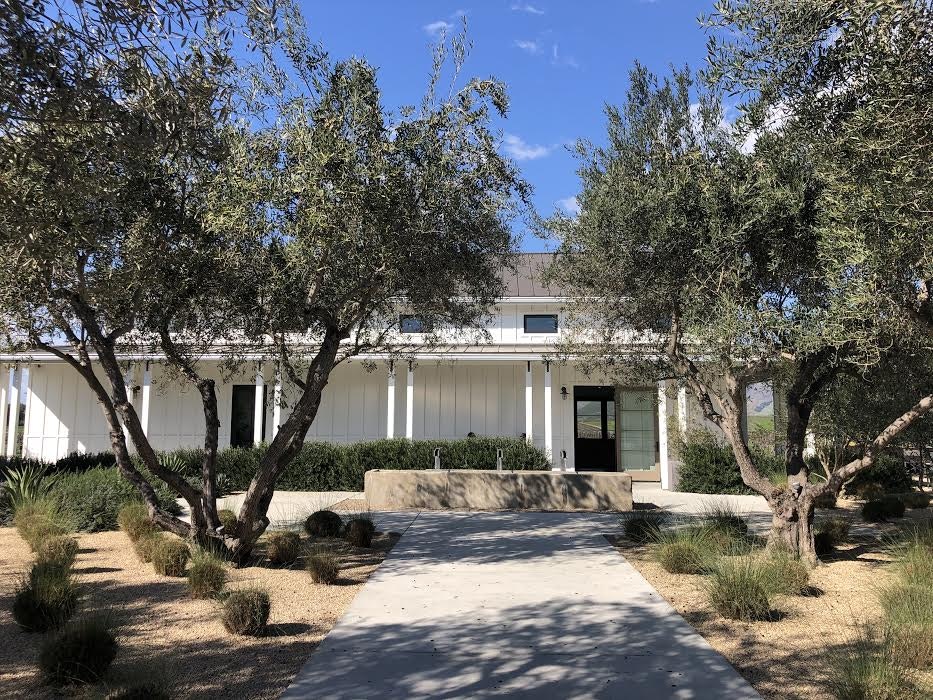 The white exterior of Biddle Ranch features a long, low porch supported by pairs of thin white columns, with a gravel walkway leading up to the structure and olive trees partially obscuring the view