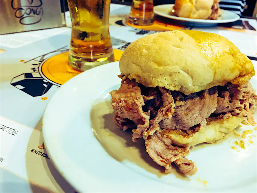 Bifana at Conga_EmilyMcAuliffeConga_Emily McAuliffe (1).jpg A close up of a pork-filled white bun sitting on a white plate, with a glass of beer in the background.