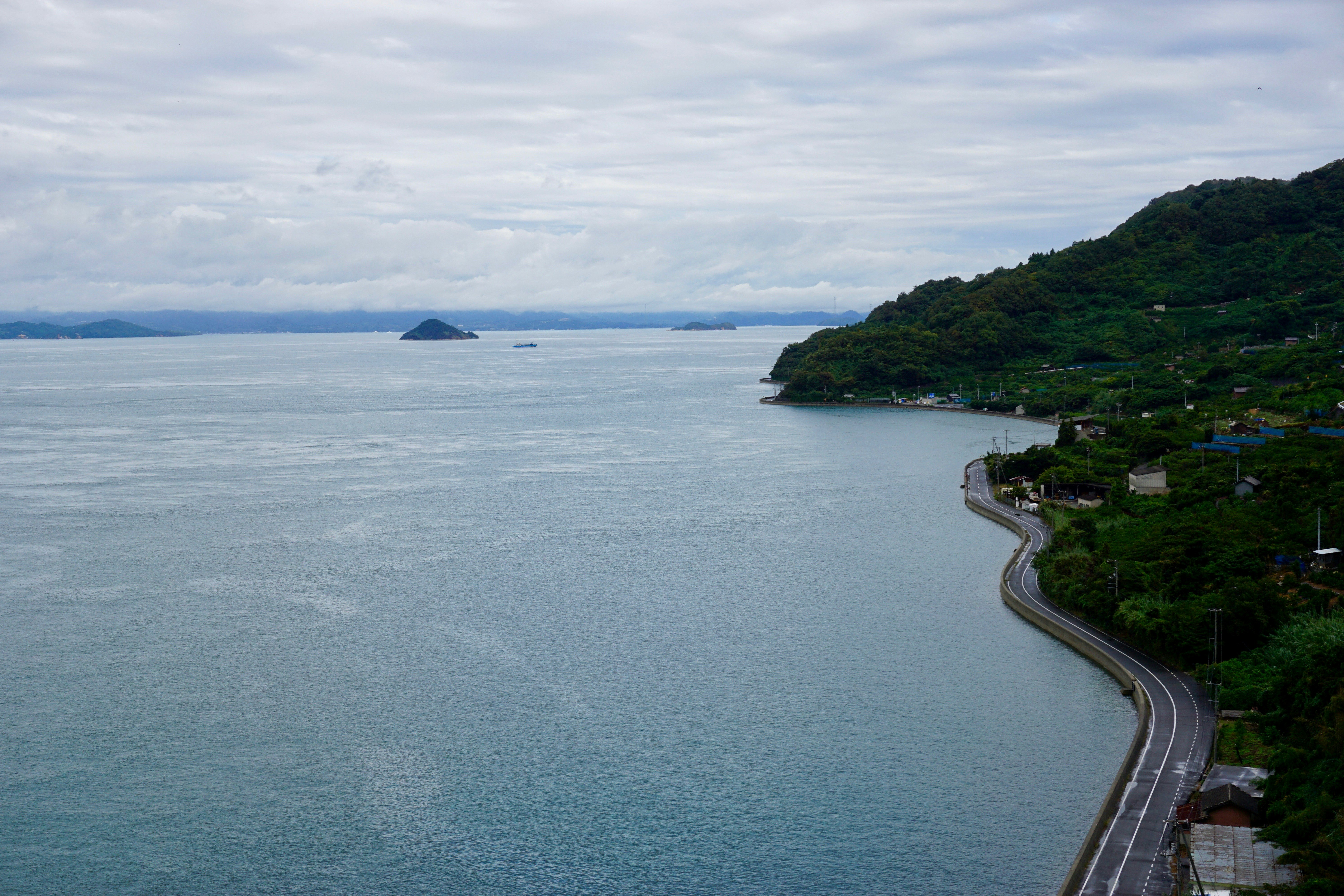 A black paved road with a white middle line highlights the winding coastline of Toyoshima. The green, thickly vegetated hills of the island come right up to the stone-washed denim sea. In the distance, a long line of low mountains follows the horizon with a white, spun-sugar cloud running parallel. A couple mounded islands stick up out of the ocean.