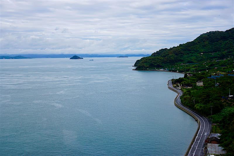 Bike-Path-Toyoshima.jpeg A black paved road with a white middle line highlights the winding coastline of Toyoshima. The green, thickly vegetated hills of the island come right up to the stone-washed denim sea. In the distance, a long line of low mountains follows the horizon with a white, spun-sugar cloud running parallel. A couple mounded islands stick up out of the ocean.