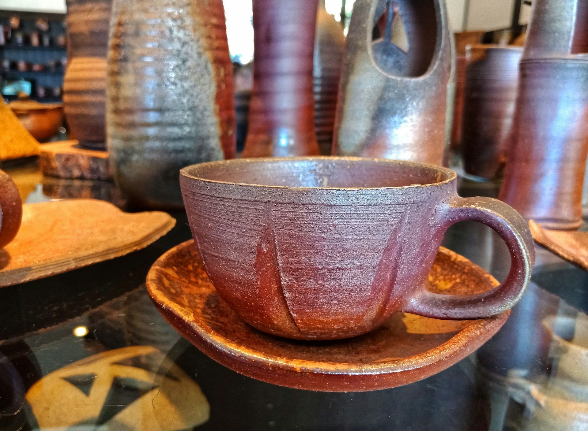 A red ceramic tea cup, made in the Bizen style, sits on a glass tabletop surrounded by other red-hued Bizen pottery.