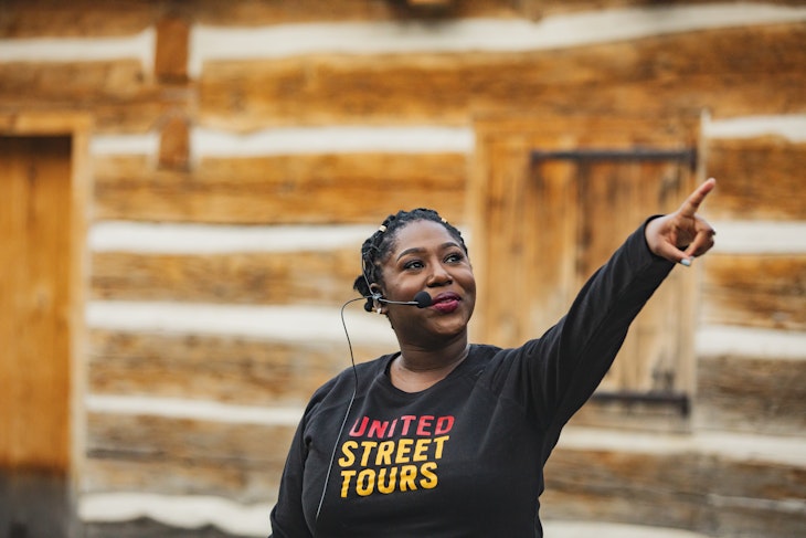 A woman wearing a head microphone and a black t-shirt that says 'United Street Tours' points to something out of frame.