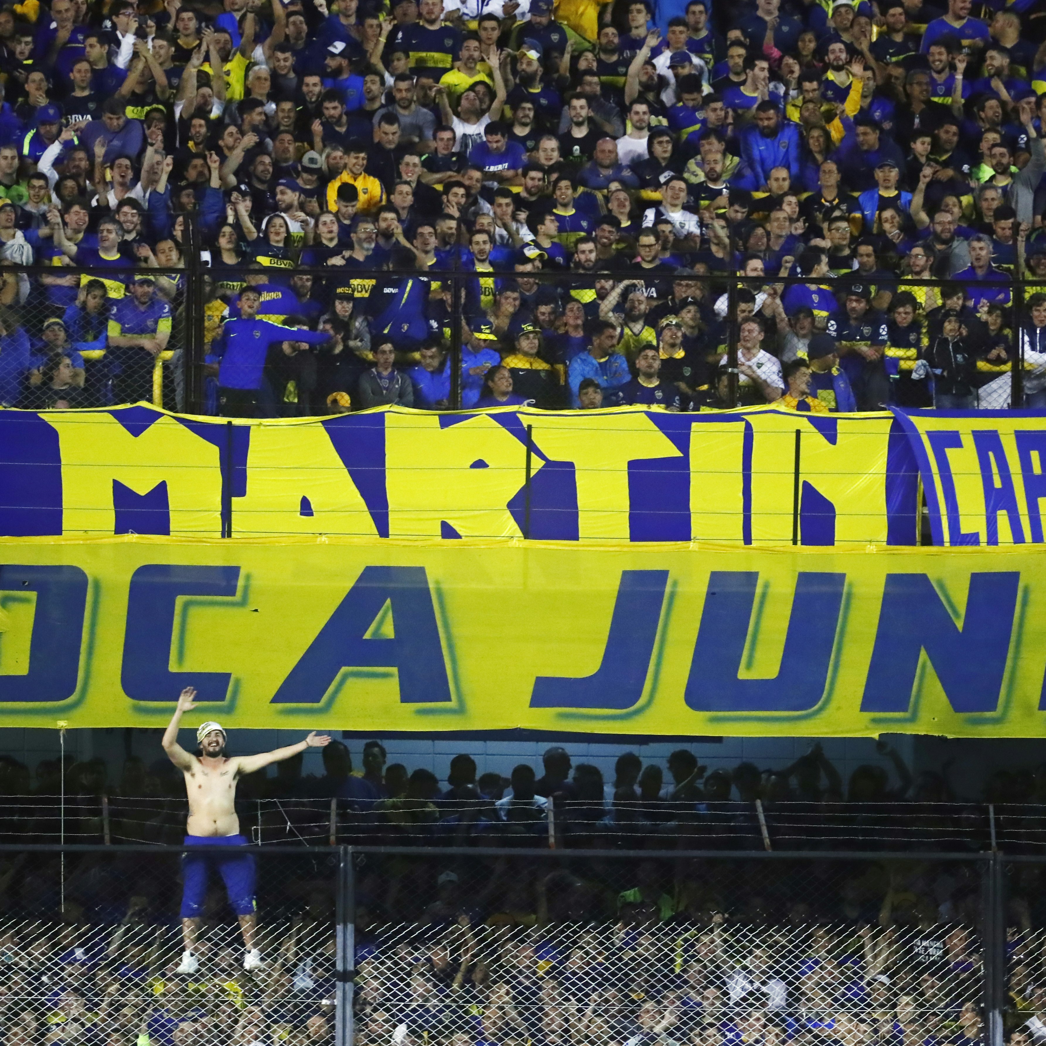 A shirtless man scales to the top of a fence topped with barbed wire at a soccer stadium in a sea of fans wearing blue and yellow in support of Boca Juniors 