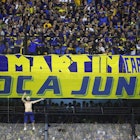 A shirtless man scales to the top of a fence topped with barbed wire at a soccer stadium in a sea of fans wearing blue and yellow in support of Boca Juniors
