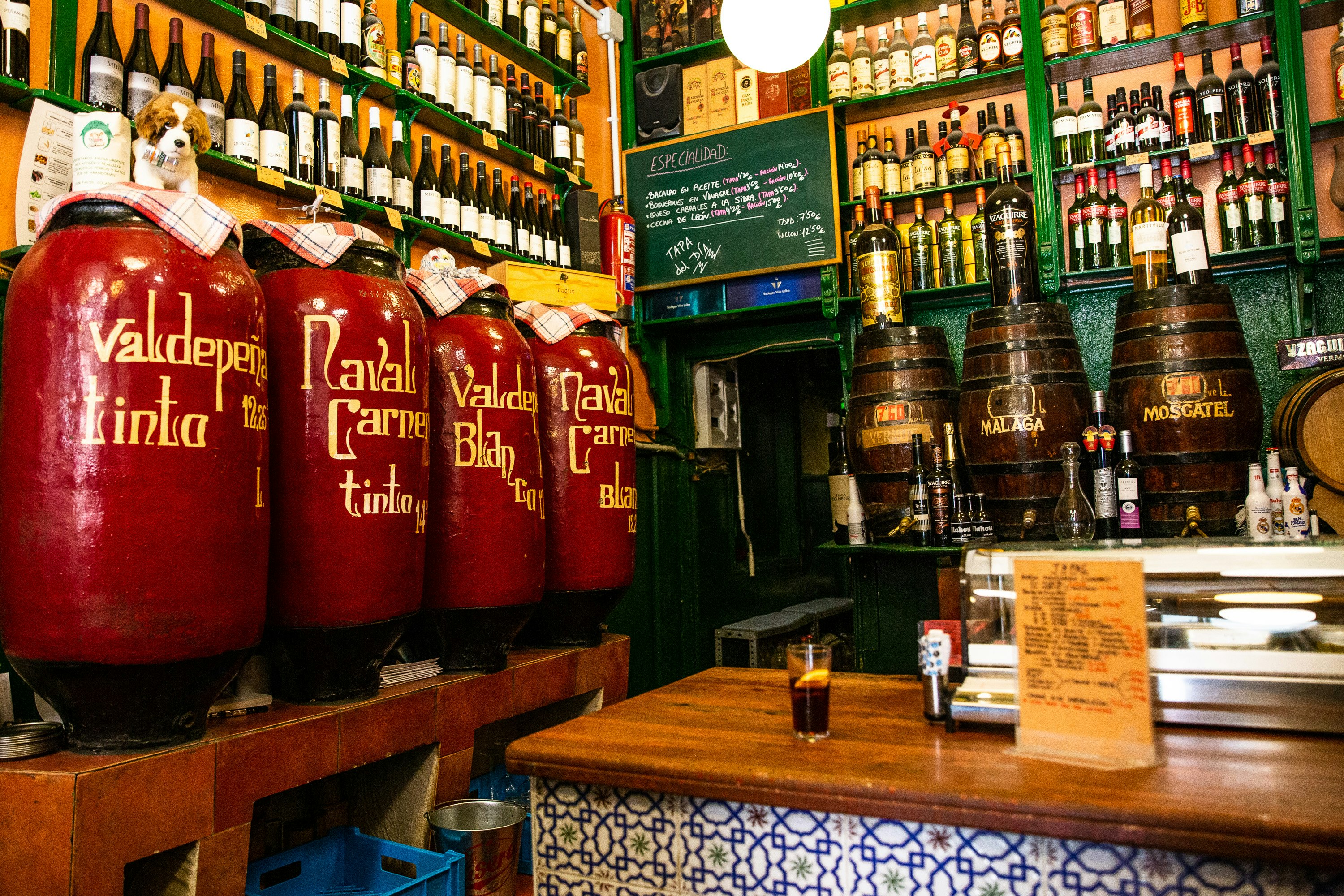 The interior of Bodegas Ricla, Madrid; the walls are lined with vats of wine, above which are green shelves packed with wine bottles.
