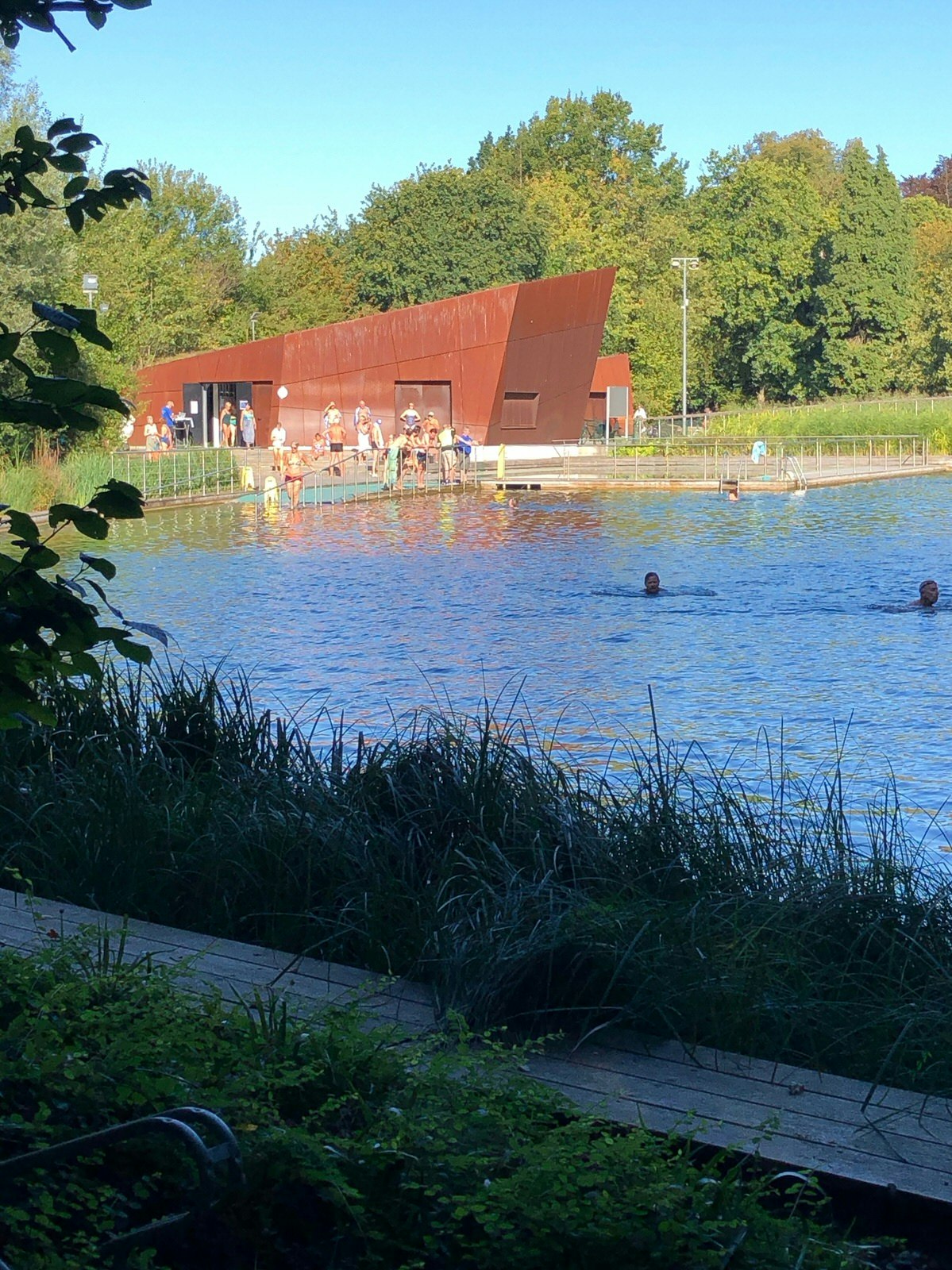 View of the natural swimming pond, Boekenger, in Antwerp on a clear sunny day. There is a small group of people swimming and lounging on the steps near the pond 