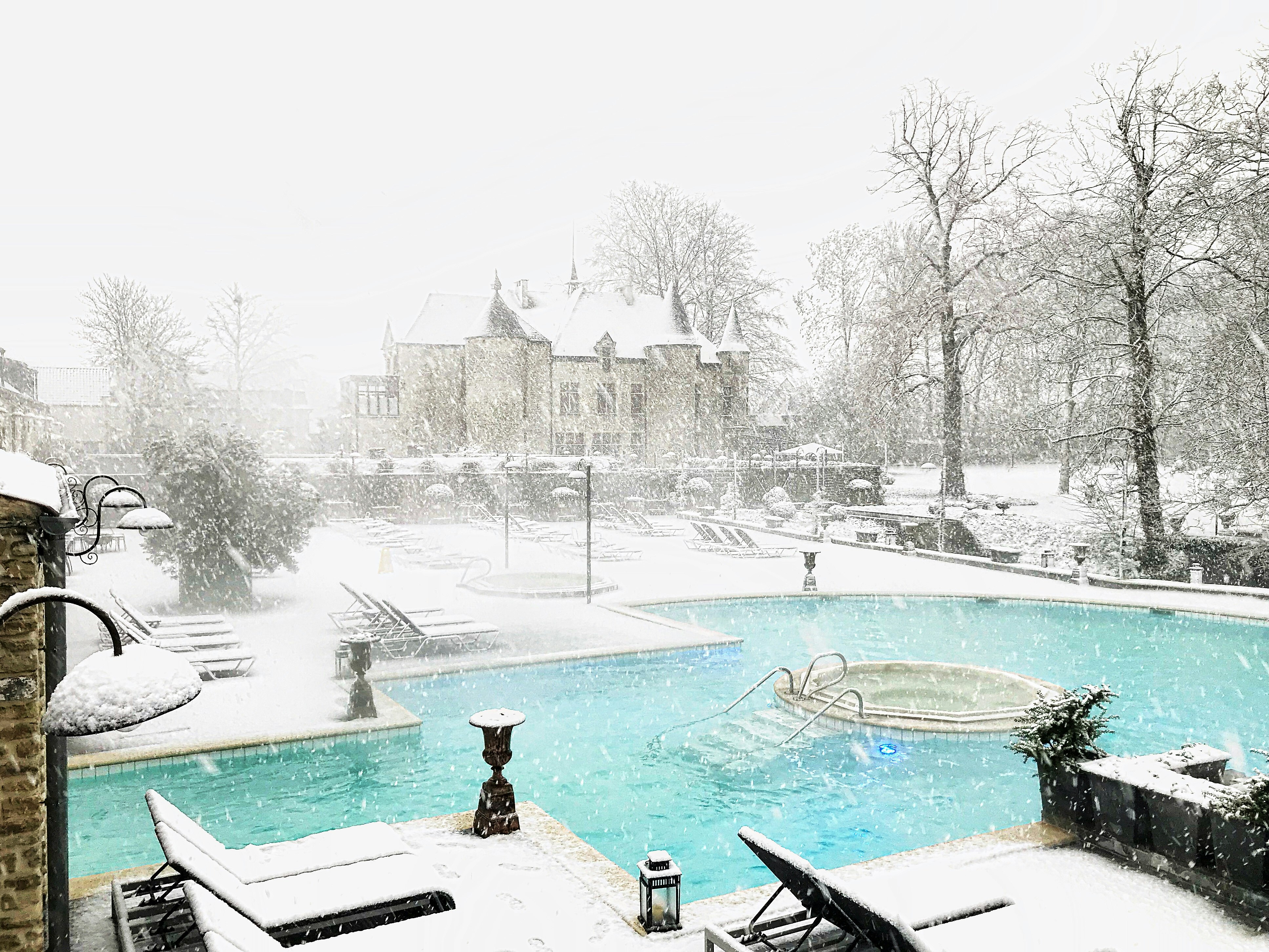 A bright blue pool with a round hot tub in the center is surrounded by snow, with a classic Belgian chalet in the background, its roof and turrets slightly obscured by fresh falling snow