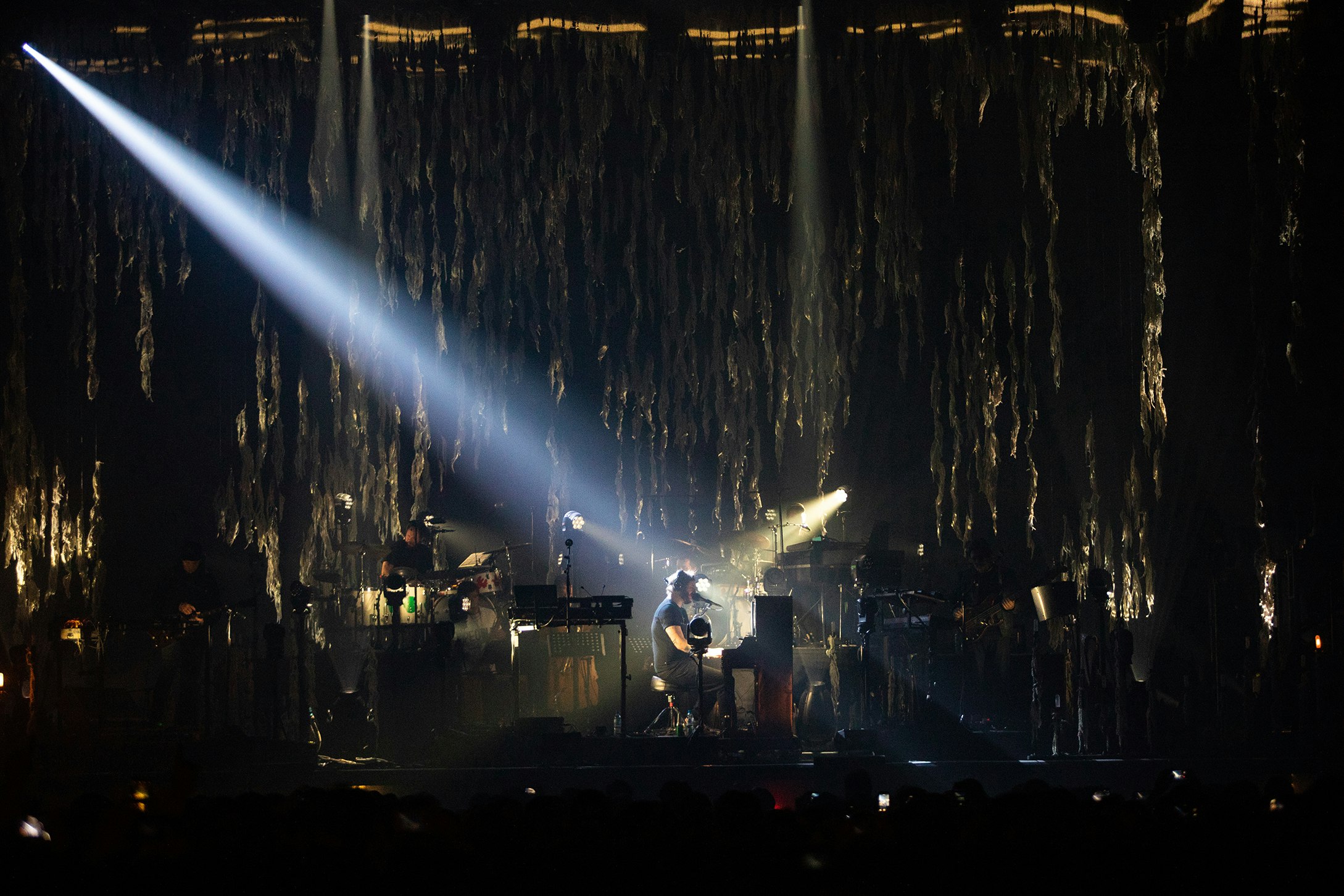 Bon Iver sitting at a piano on a very dimly lit stage in front of a large crowd at Pitchfork Music Festival in Paris, France. 