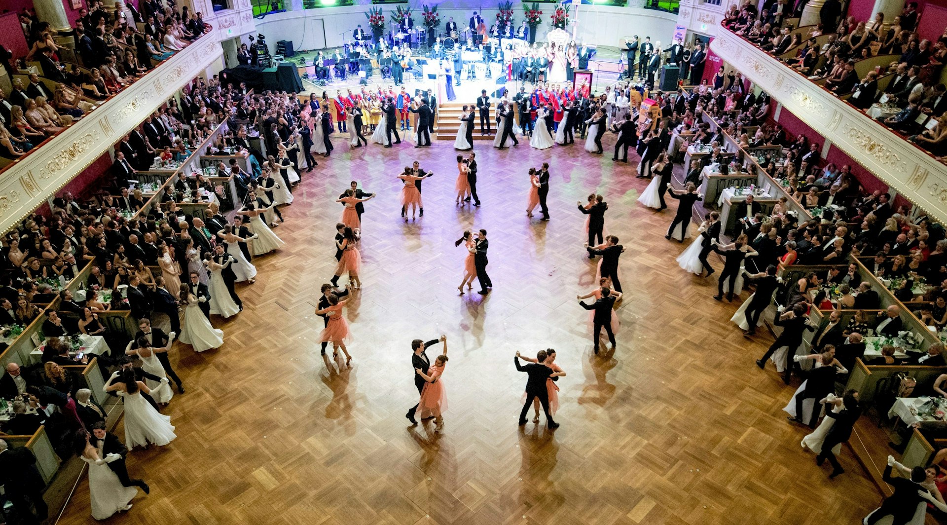 Couples fill the centre of the dance floor, with men in dark suits and women wearing peach-coloured dresses. A live band can be seen at the far end of the dance floor