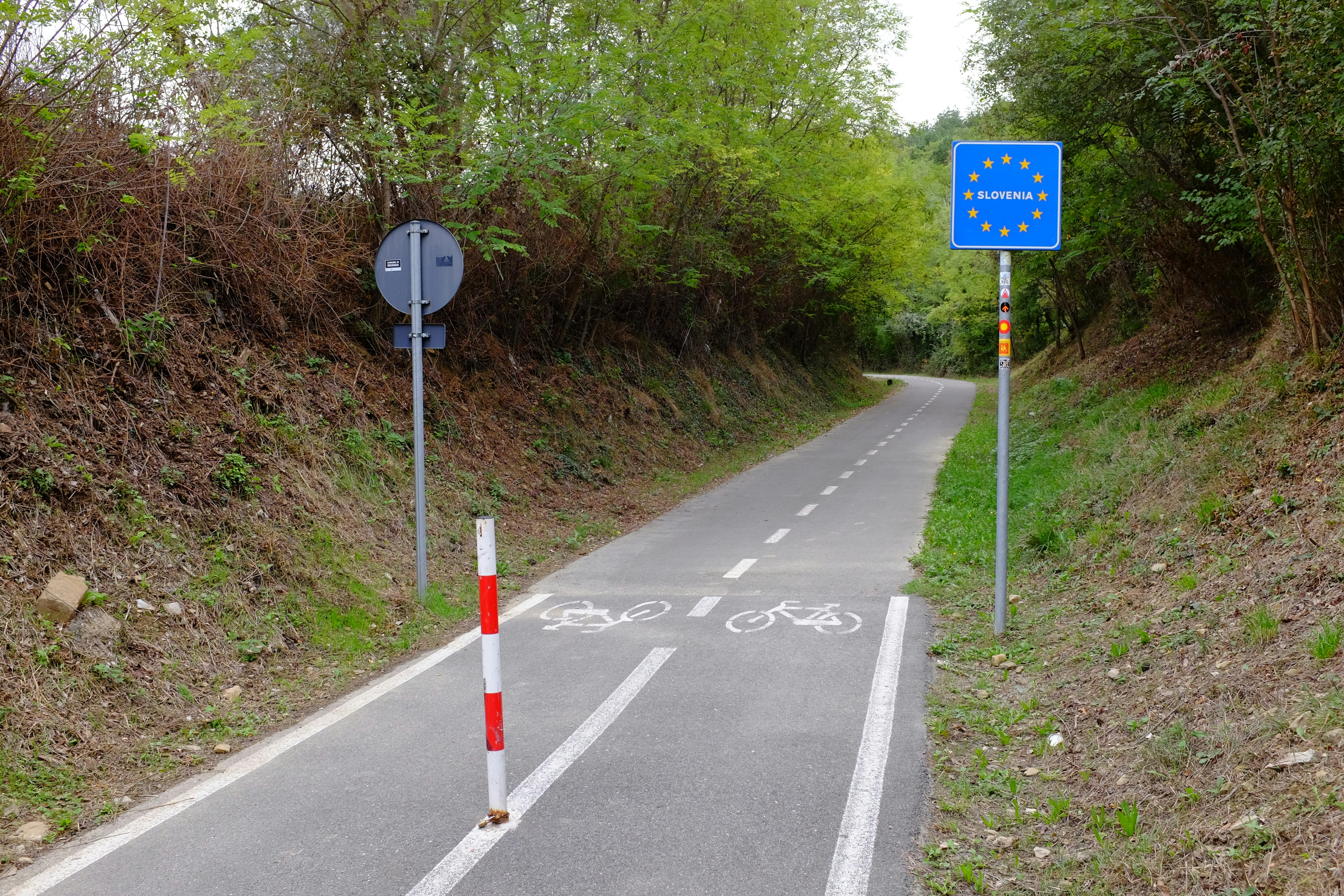 The border between Italy and Slovenia on the Parenzana Trail, which are two narrow lanes marked by white lines, bicycle traffic logos and  a blue sign facing the viewer with the blue field and yellow stars of the European Union