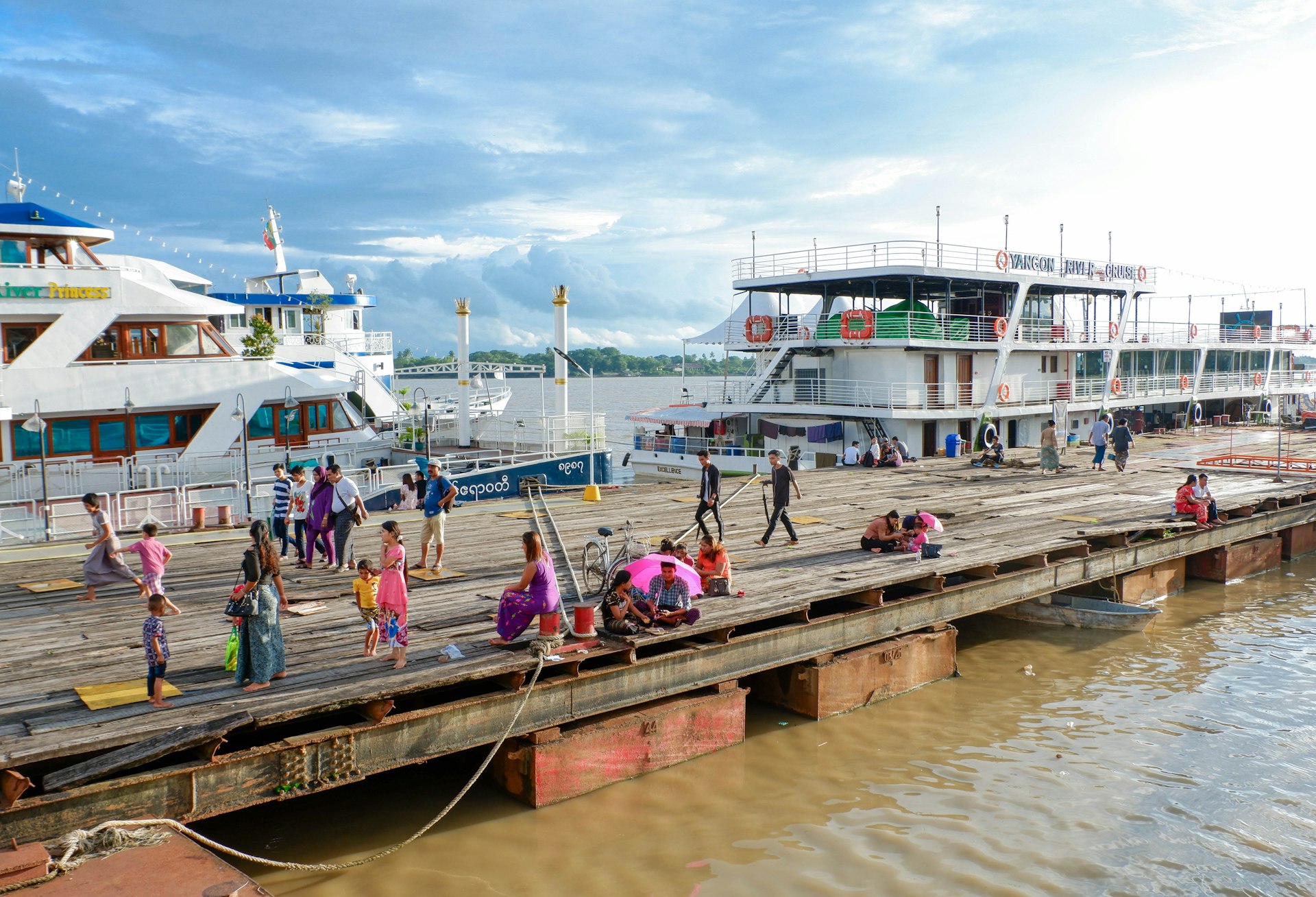 Groups of people sit and walk along wooden Botataung Jetty, which splinters out into Yangon River. In the background a couple of large yachts are visible.