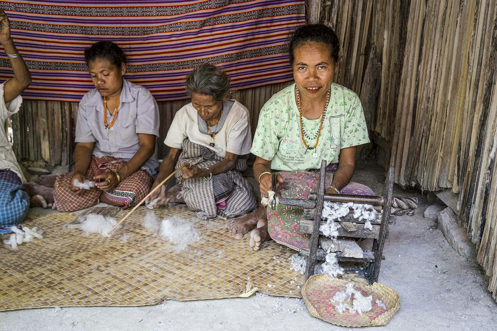 Three women sitting on a woven mat and dressed in pastel clothing work with fibers to make string. Boti, East Nusa Tenggara, Indonesia.