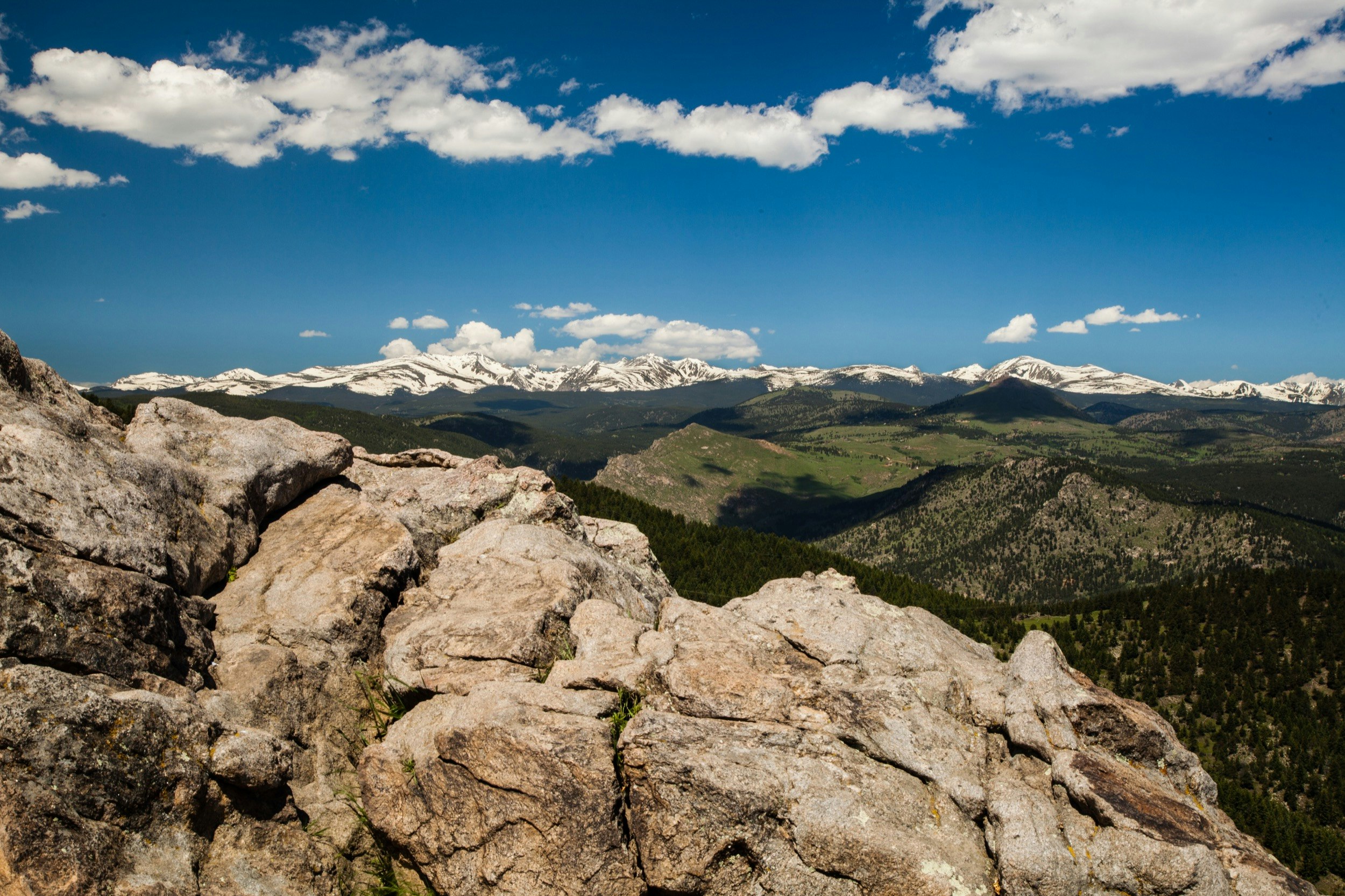 Snow capped mountains rise in the distance beneath a brilliant blue sky as large boulders and rocks dominate the foreground of a mountain near Boulder, Colorado; Hiking
