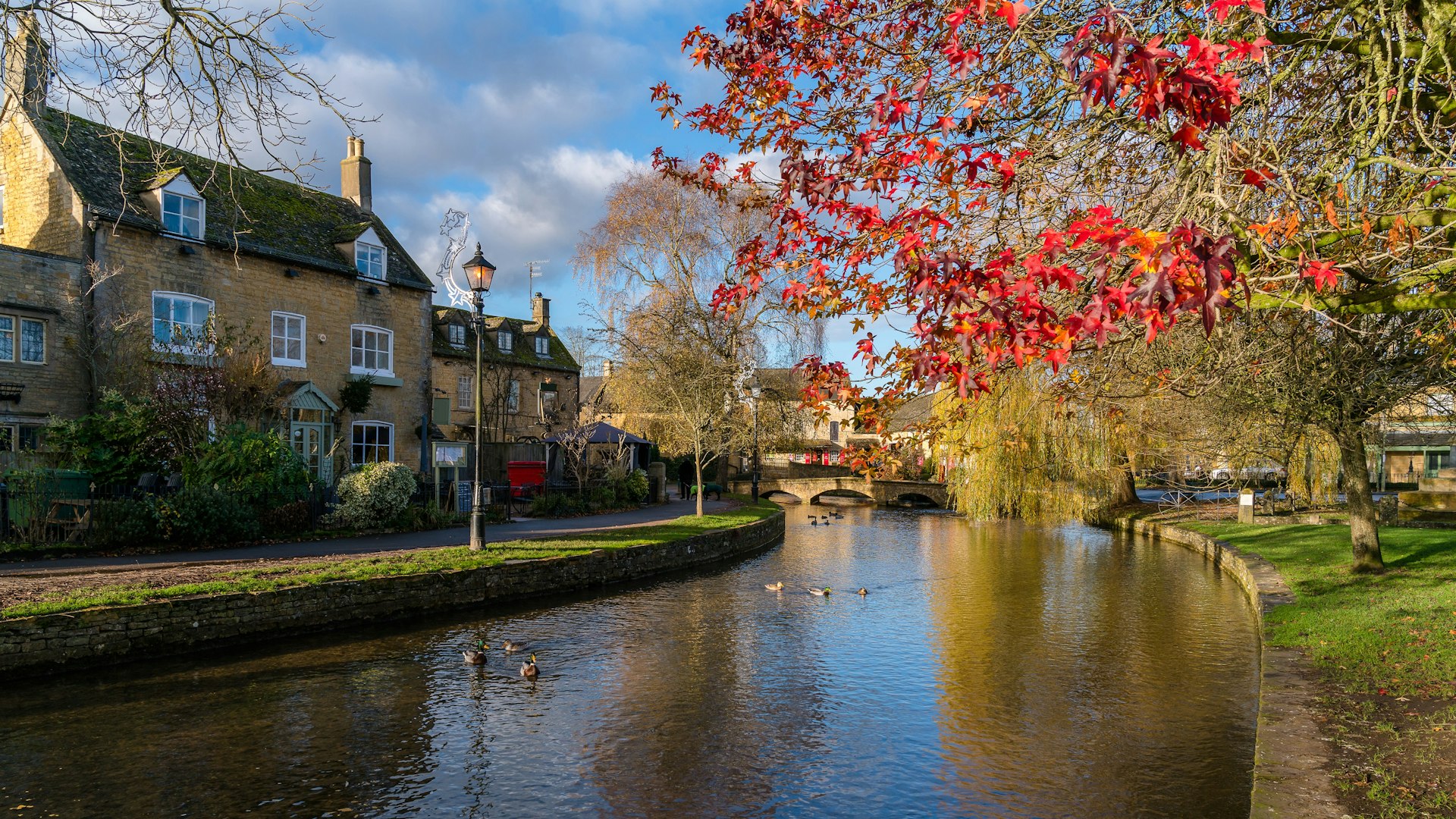 A river meanders past a honey-coloured stone house on one side and a tree bright with red autumnal; leaves on the other.