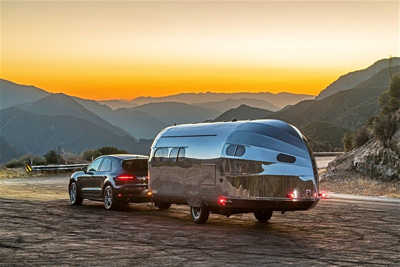 A silver luxury trailer and vehicle sit in front of a sunset and mountain background. 