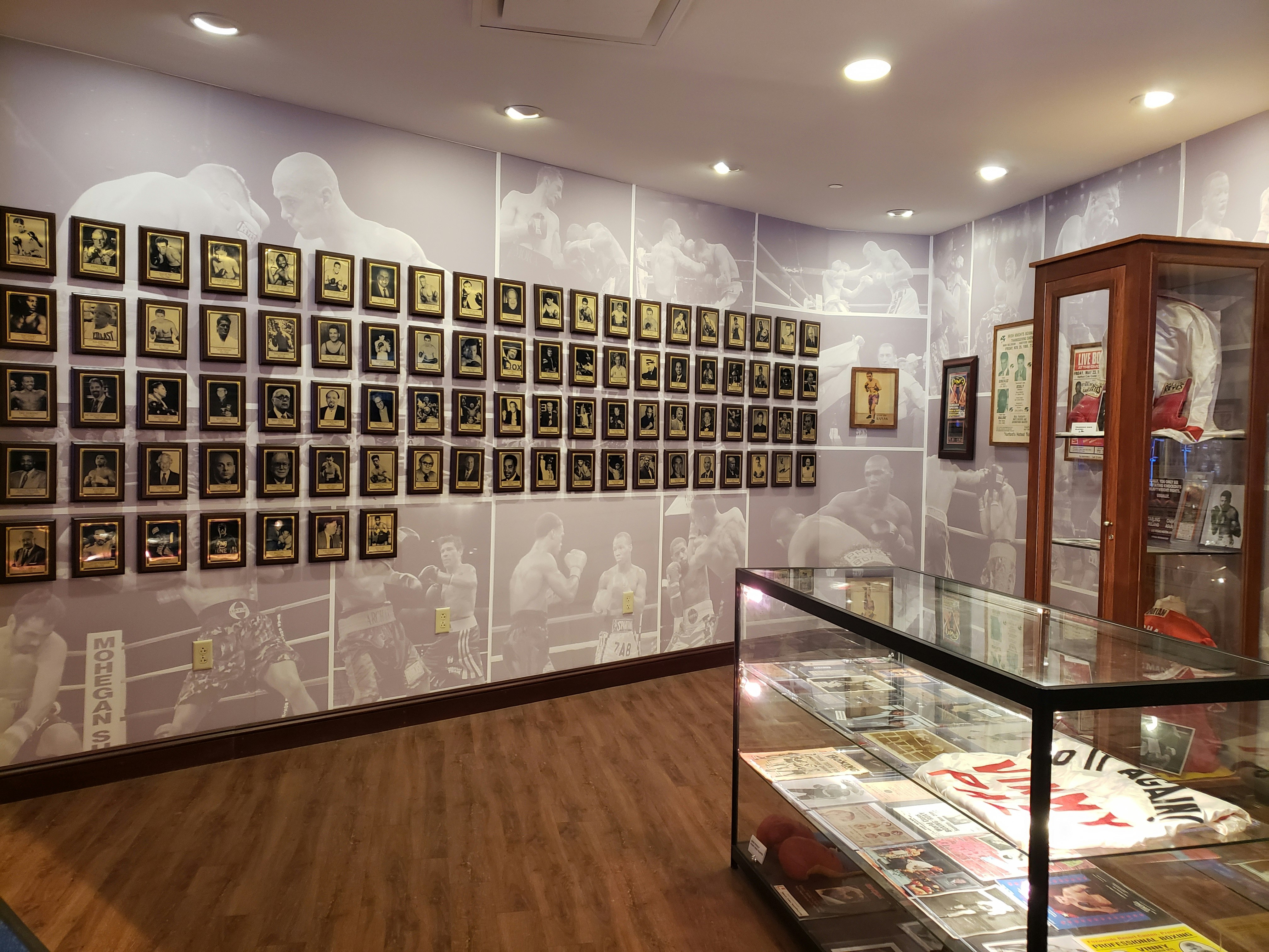 A curved wall at the Connecticut Boxing Hall of Fame is taken up with wooden plaques, on which sepia tone photographic portraits of great boxing champions are mounted. To the right are two glass display cases full of memorabilia. 