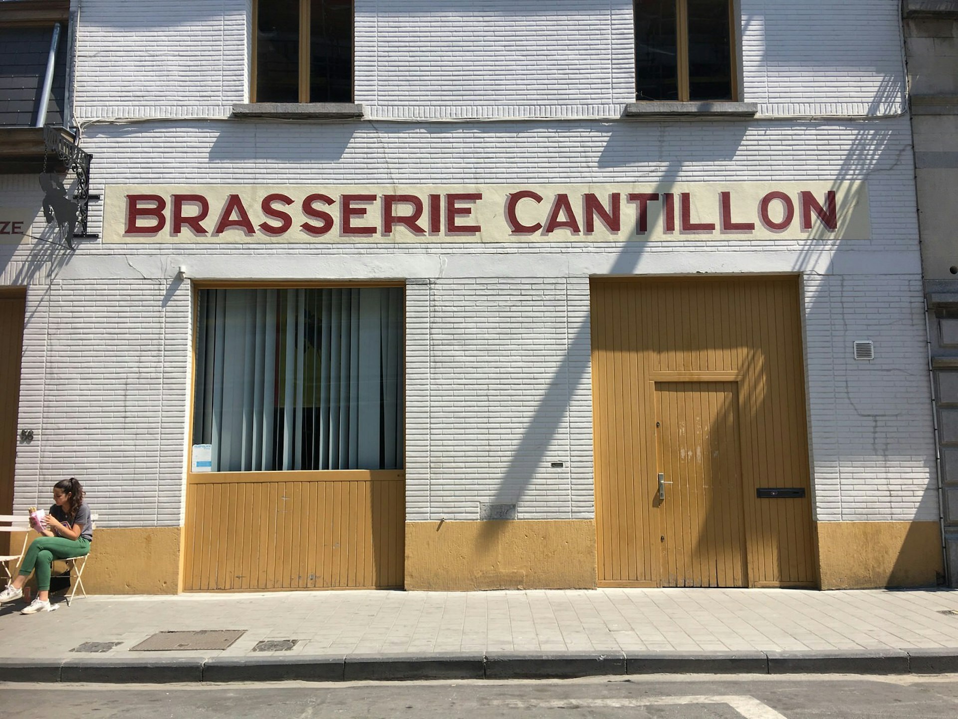 Brasserie Cantillon facade - A two-storey building with large square windows. The bottom of the building is painted a dusty yellow and 'BRASSERIE CANTILLON' is painted in capital red letters above the yellow door. A woman is sitting at a table outside having a beer. 