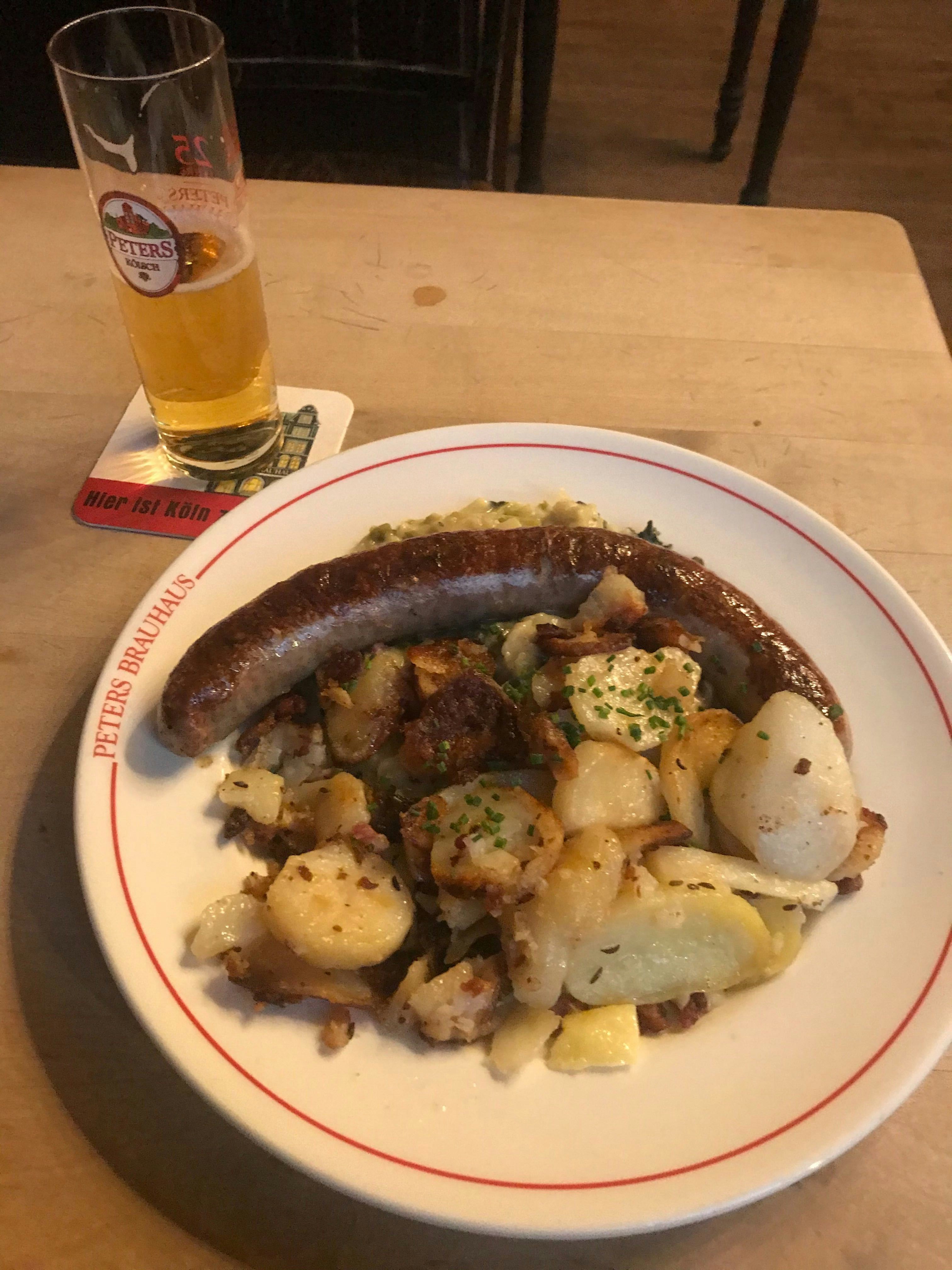 A sausage and fried potato on a plate with a small beer next to it.