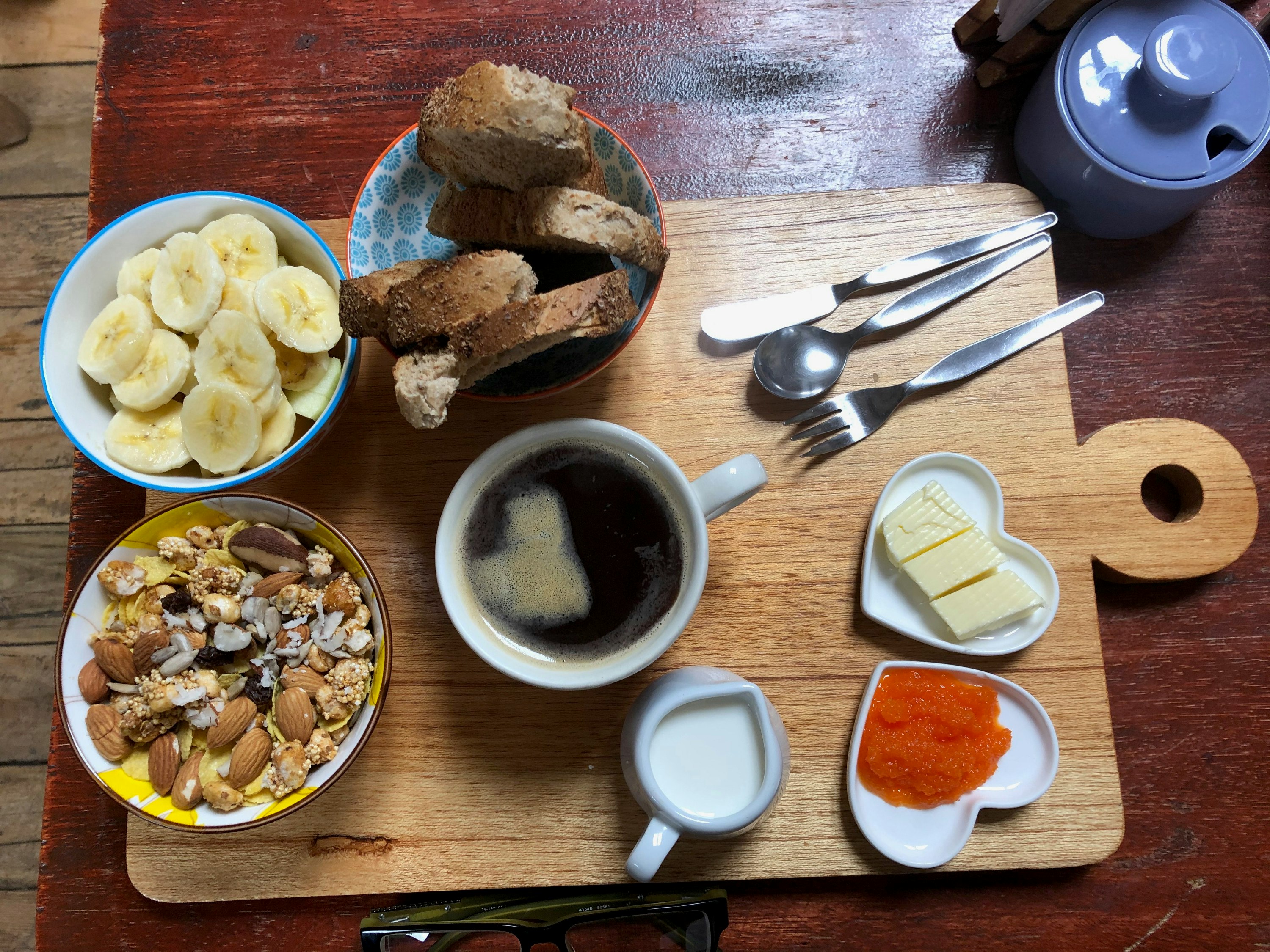 Bananas, bread, coffee, granola, butter and jam on a wooden slate.