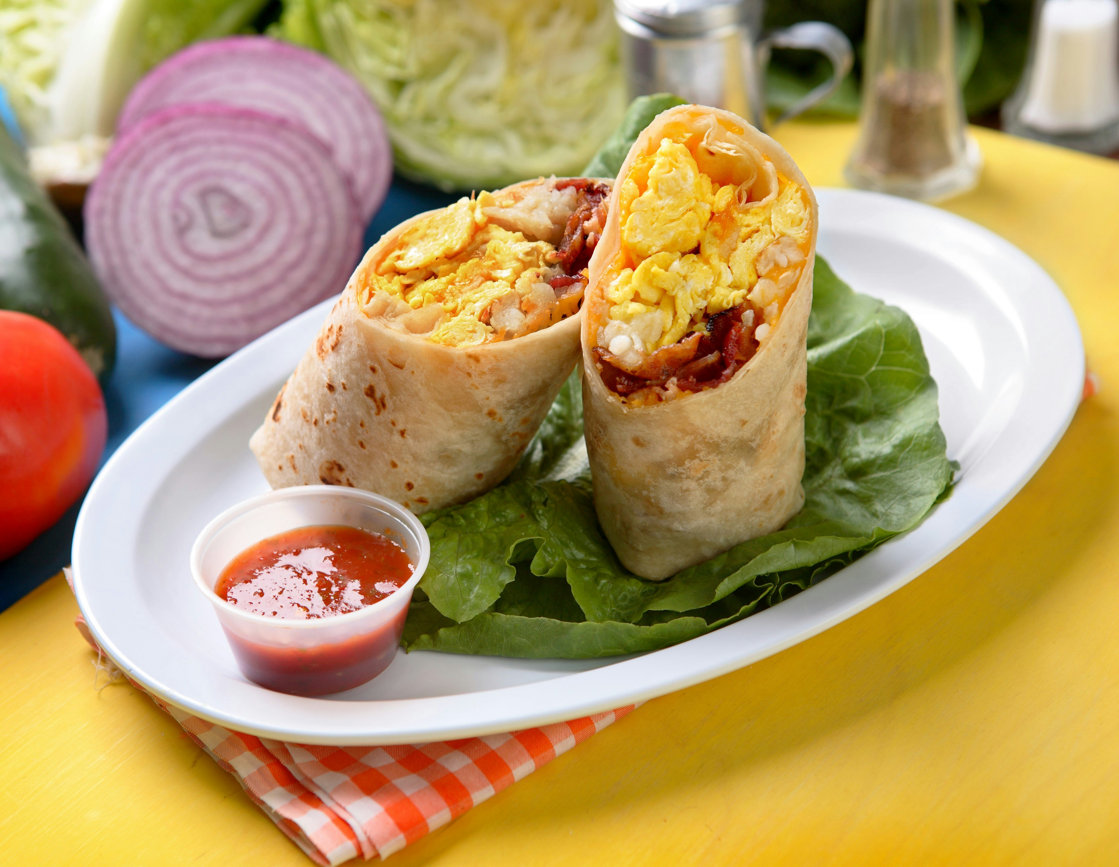 A breakfast burrito that has been sliced in half, sits on a plate on a tabletop. The burrito, contains egg and bacon, wrapped inside a tortilla.