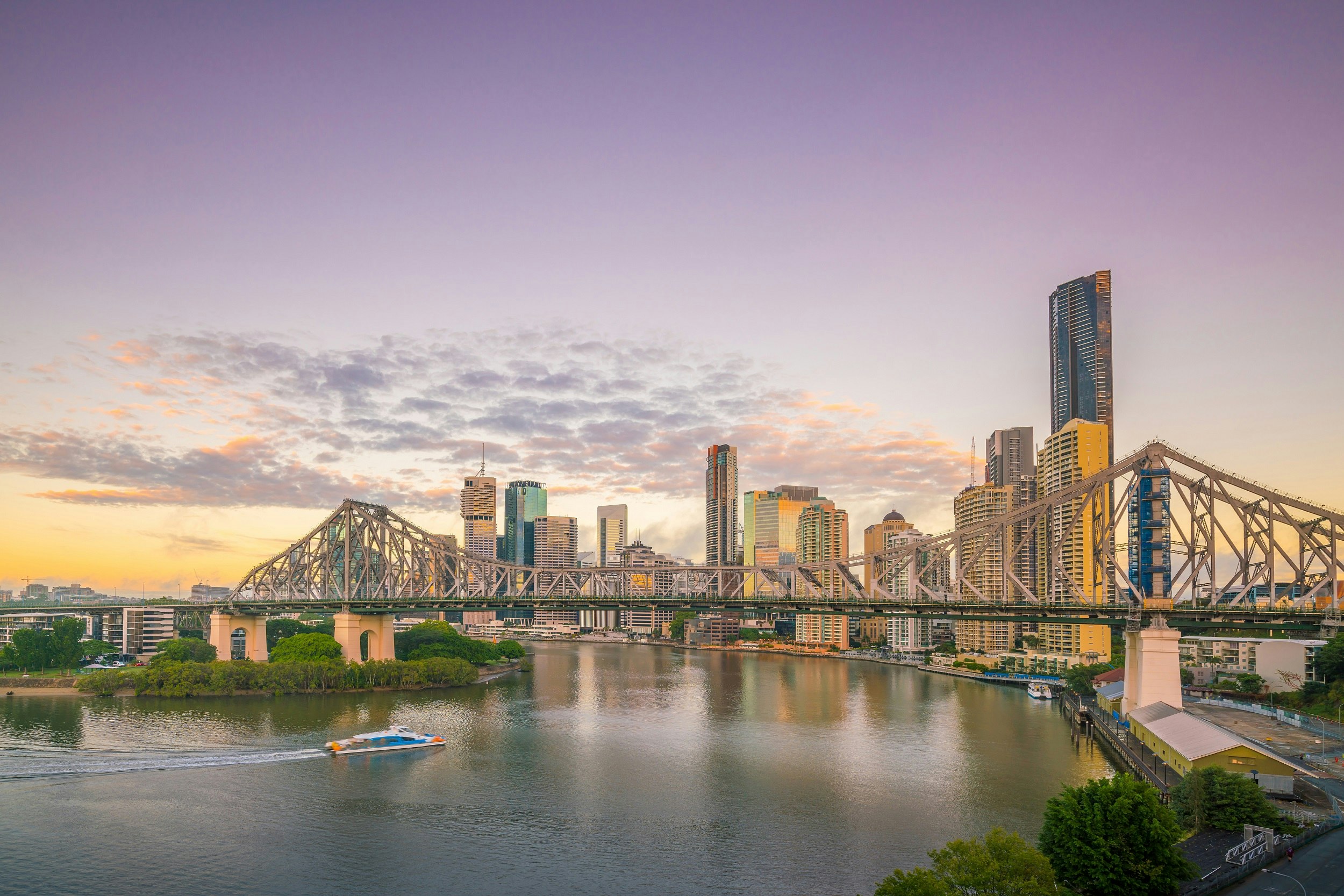 A skyline shot of Brisbane at sunset with a purple sky; the scene looks over a river and large rail bridge, with the cityscape as a backdrop.