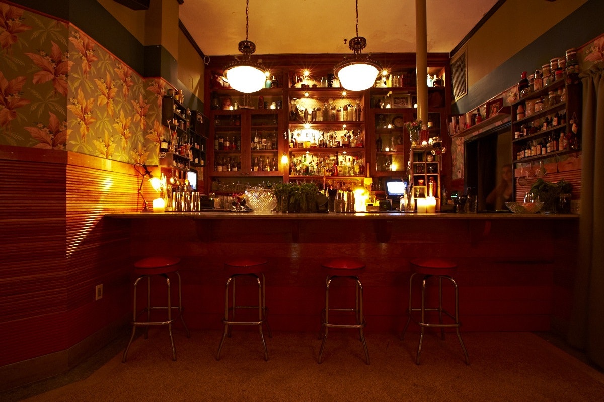 Inside a seductively lit cocktail bar with vintage decor and pendant lights suspended from the ceiling. Behind the bar there is a wooden cabinet stocked with bottles and ingredients.