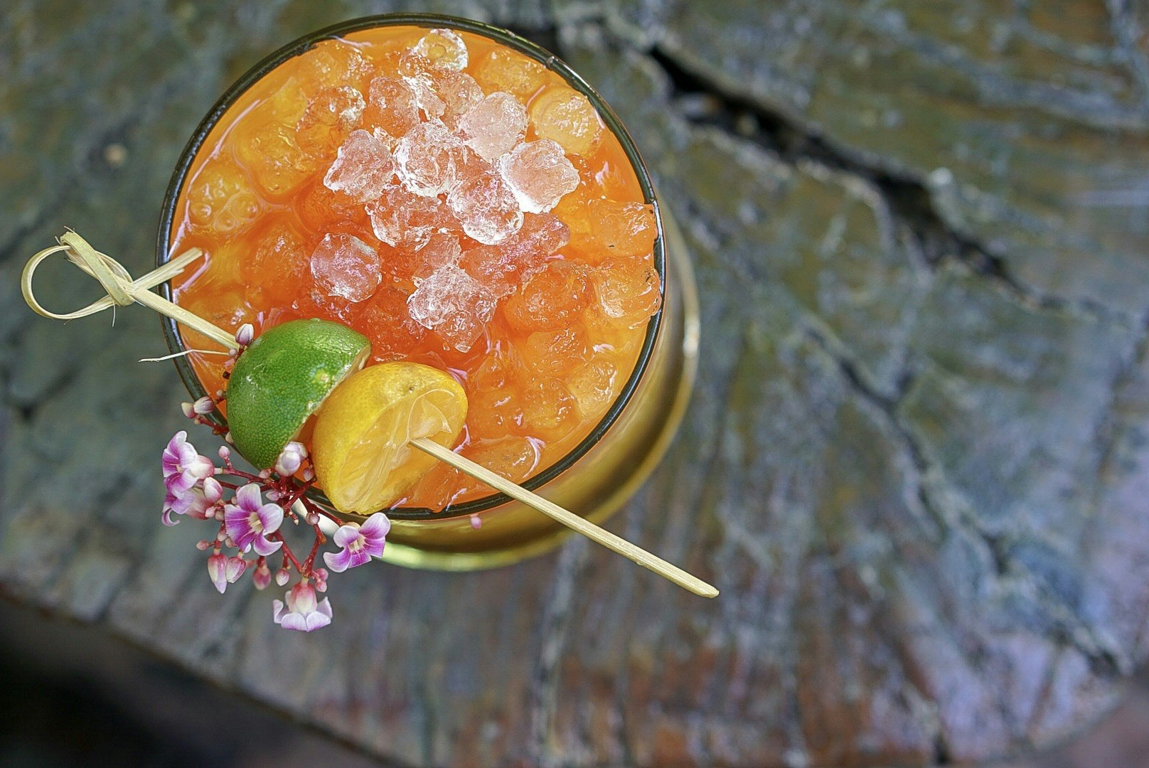 View looking directly down upon a glass containing a bright orange cocktail. The drink is muddled with crushed ice, and a wooden skewer with green and yellow fruits rests artfully across the rim, as well as a delicate sprig of flowers.