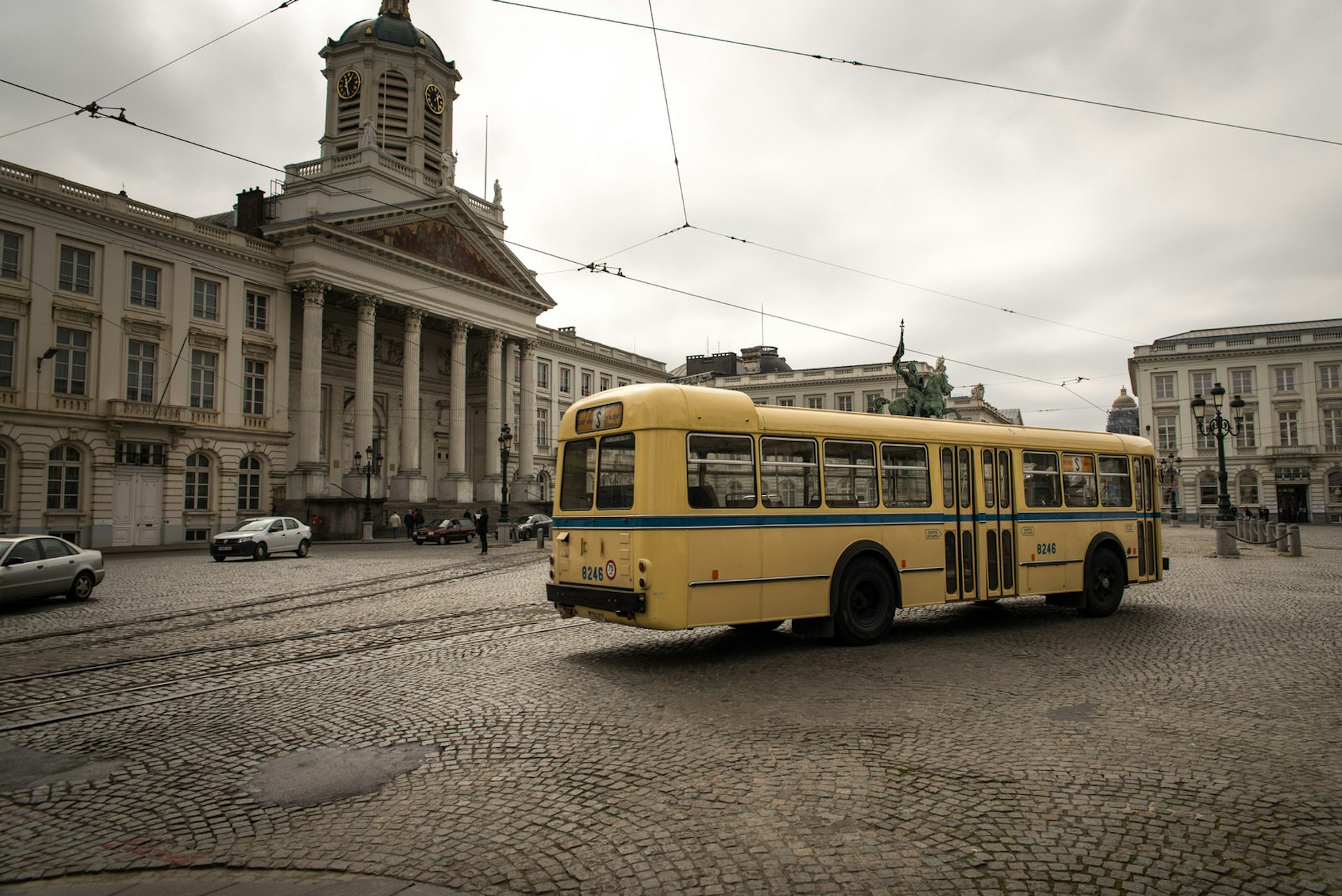 An old-fashioned yellow school bus is parked in a large cobbled square in Brussels with an elegant columned-building in the background on an overcast day