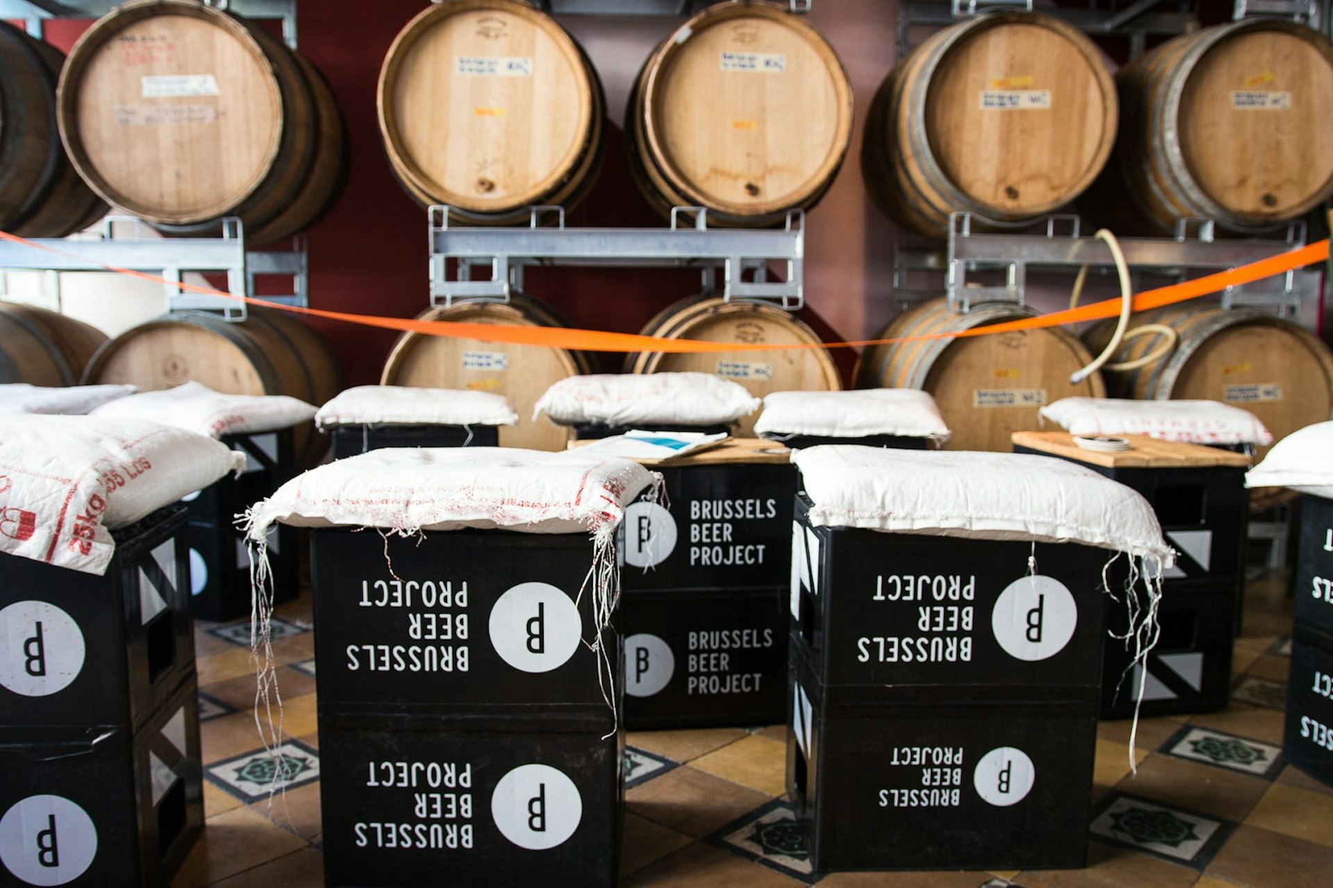 black plastic beer crates with 'Brussels beer project' and the company logo printed on them are stacked in front of wooden barrels. Sacks are placed on top of the crates to make seats. 