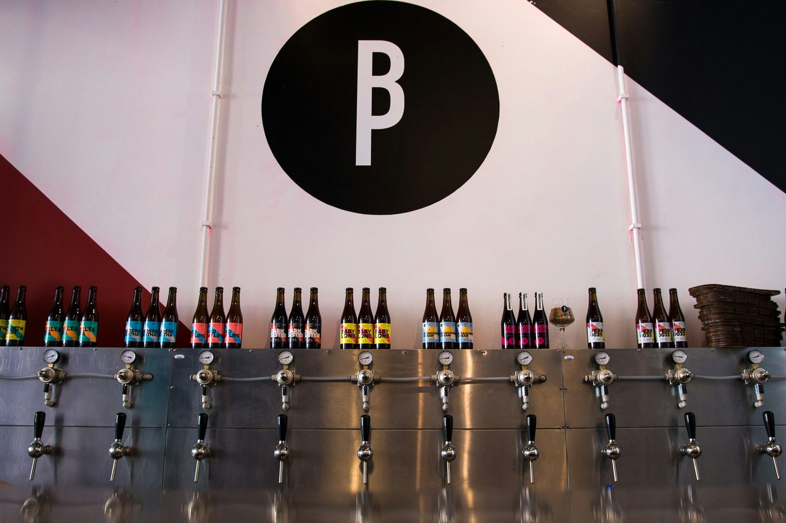 A steel bar with several beer taps with black plastic handles. Bottles of beer with colourful labels have been placed above each tap to show which beer is on tap. There is a large black logo of a capital B and P painted on to the white wall. 