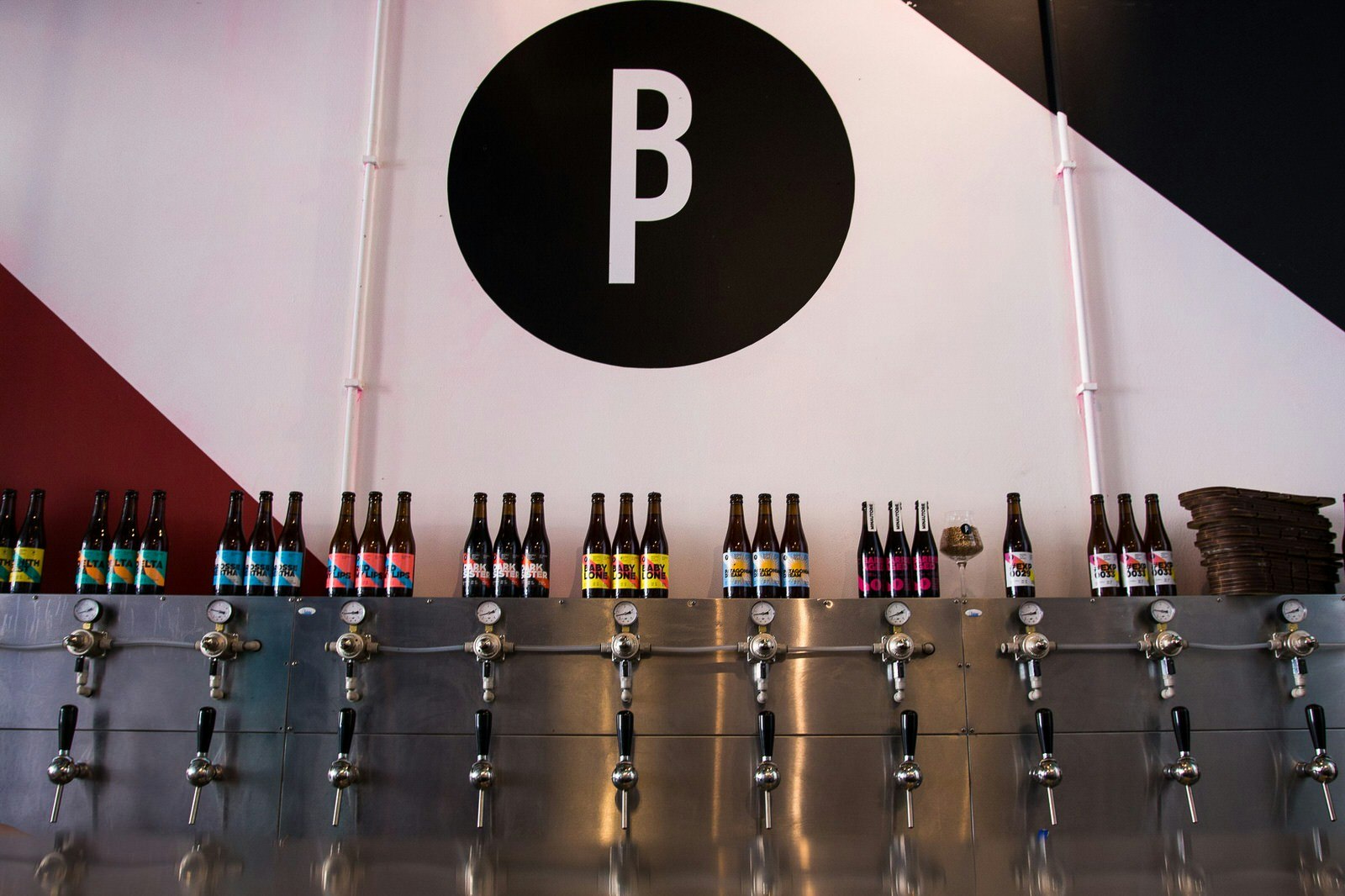 A steel bar with several beer taps with black plastic handles. Bottles of beer with colourful labels have been placed above each tap to show which beer is on tap. There is a large black logo of a capital B and P painted on to the white wall.