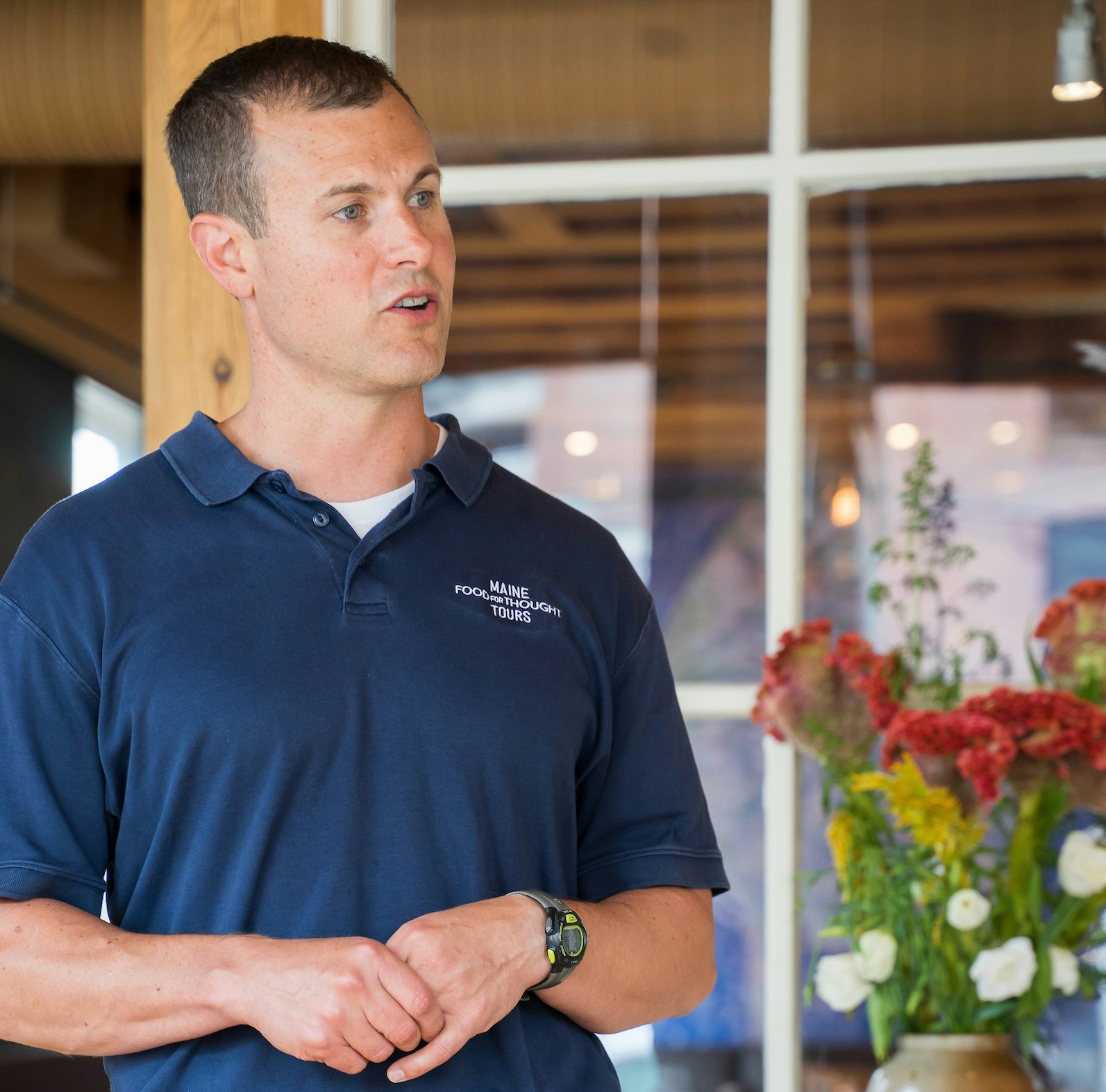 Bryce Hatch, co-founder of Food for Thought Tours, stands by a vase of red and yellow flowers in a blue polo shirt with the name of the business. He looks to his left.