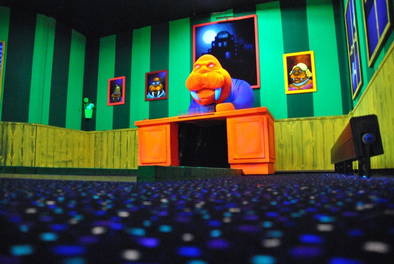 A large, glow-in-the-dark model of a walrus in a suit, sitting behind a desk, forms part of the course at Bubbel Jungle Golf in the Netherlands. The walls behind the model are painted in striped green, giving the whole scene a psychedelic effect.