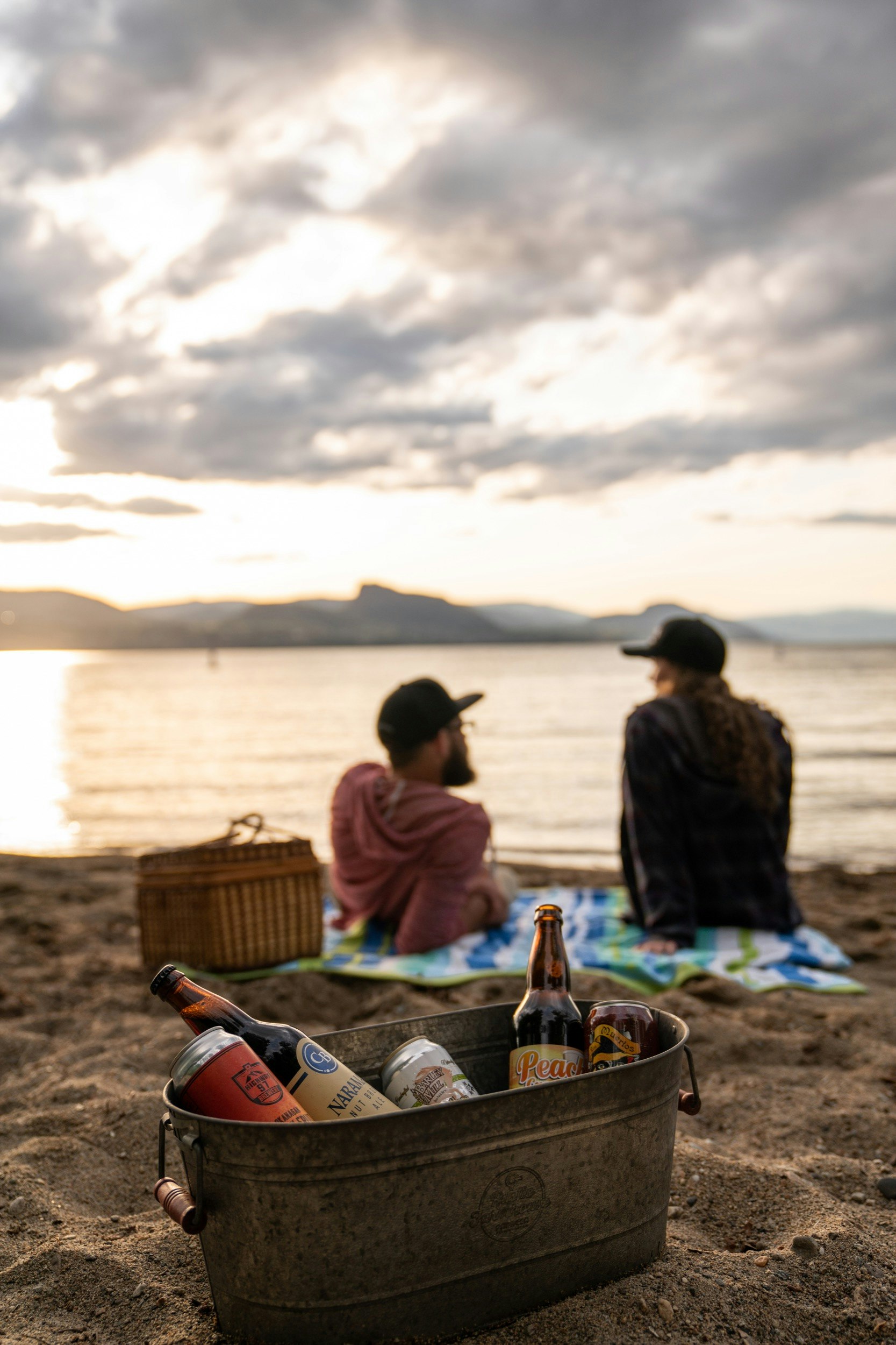 En metallhink med ett urval av hantverksöl sitter bakom ett par och picknickar på sanden bredvid en sjö.